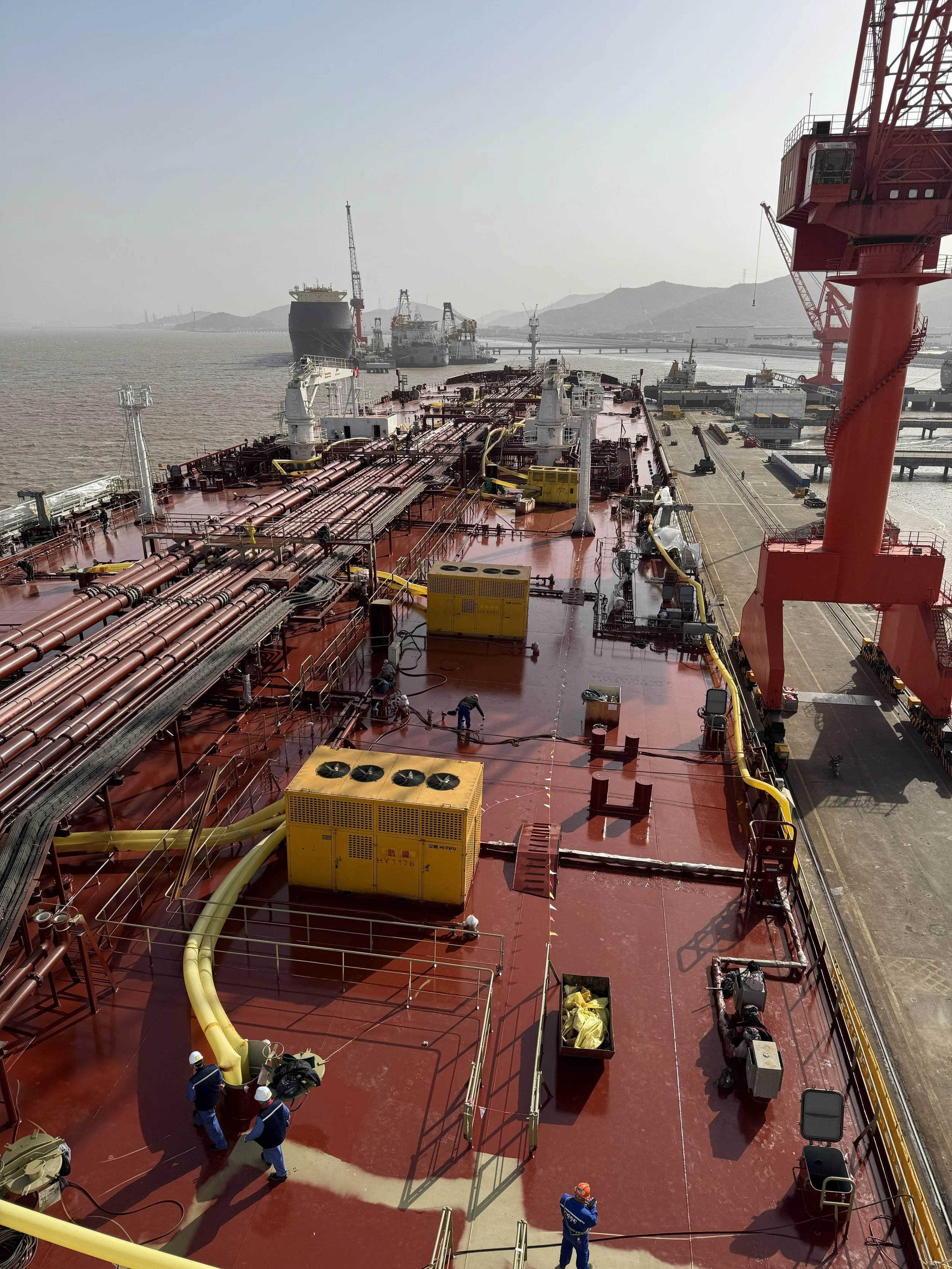 View of large ship deck with red surface, yellow pipes, cranes, and workers wearing safety helmets.