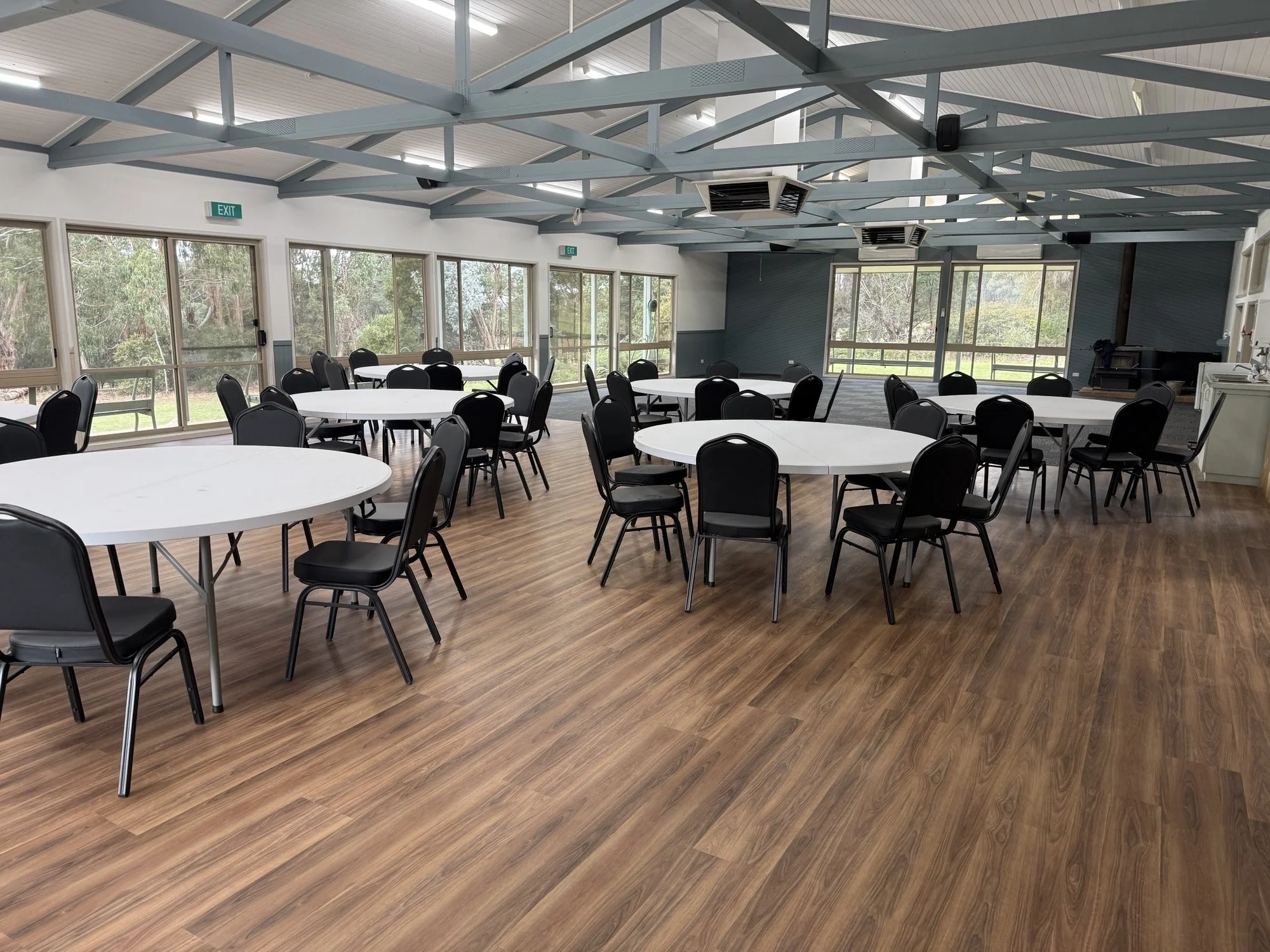 An indoor event space with round tables and black chairs, large windows showing trees outside, a wooden floor, and a high ceiling with metal beams and lighting.