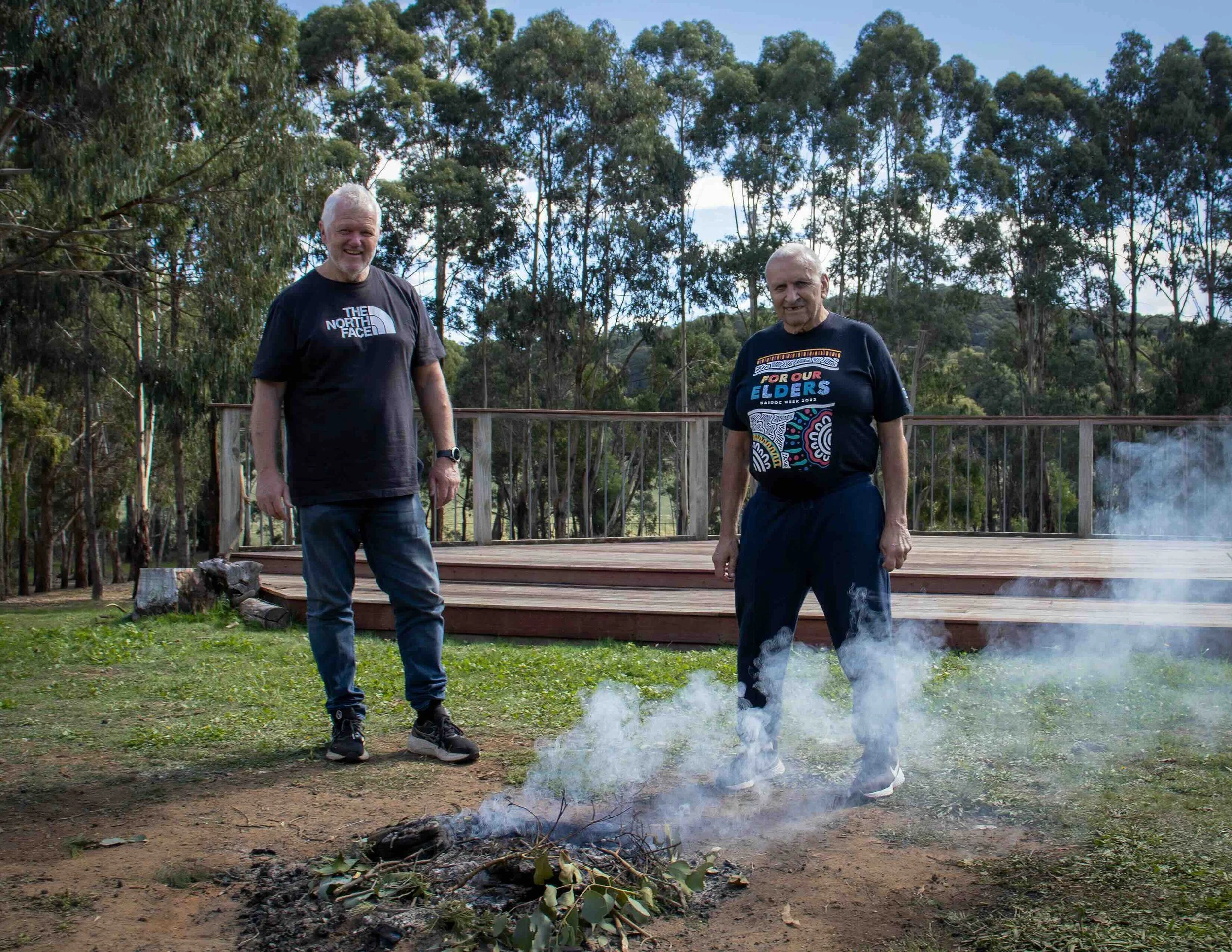 Two elderly men standing outdoors on a wooden deck, with trees in the background. One man is wearing a black t-shirt and jeans, the other a black T-shirt with colorful patterns and the words 'For Our Elders,' and both are smiling. Smoke rises from a small fire on the ground.