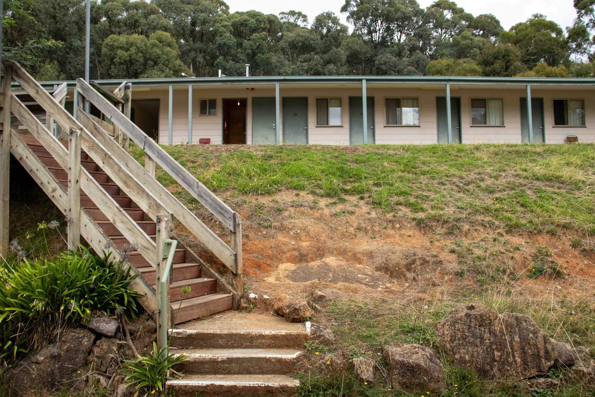 Wooden staircase leading up a grassy hillside to a low, single-story building with multiple doors and windows, surrounded by trees.