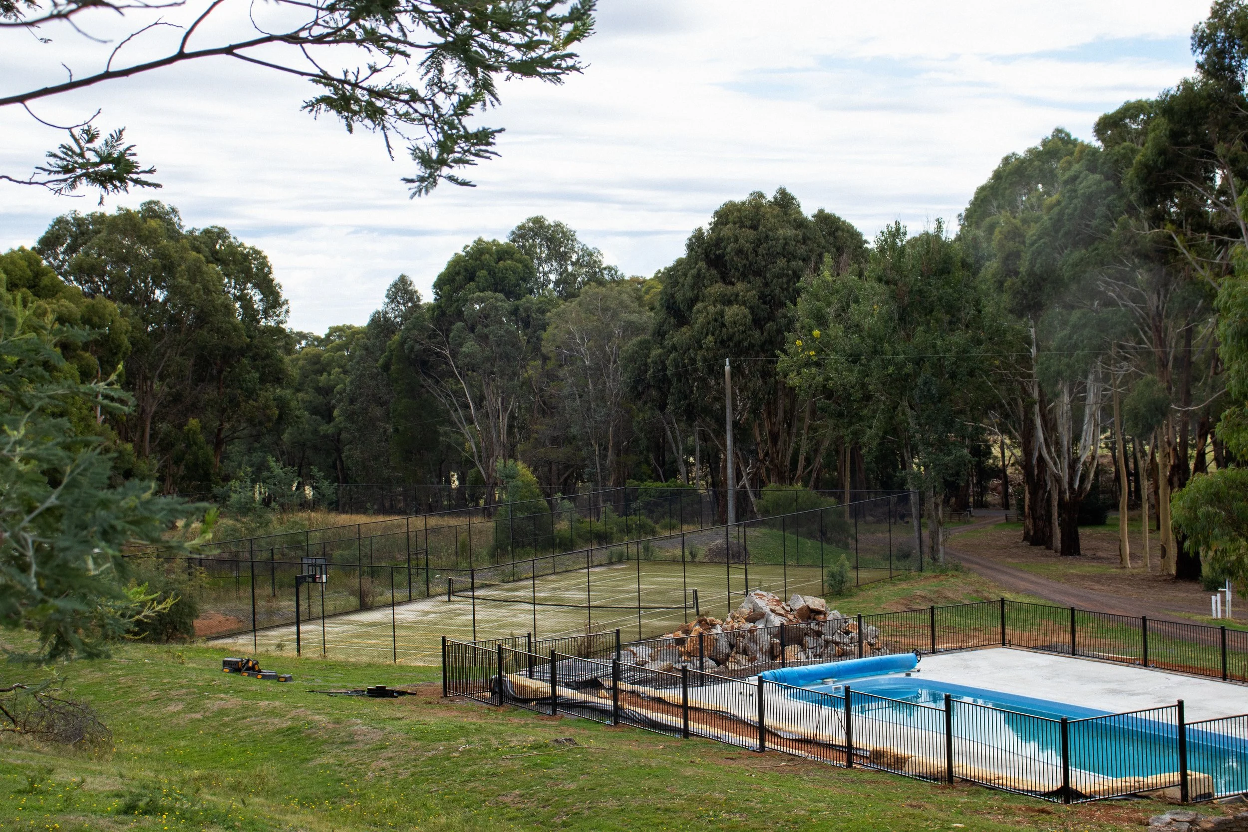 Backyard with a tennis or basketball court, a small swimming pool, and a wooded area with tall trees and a cloudy sky.