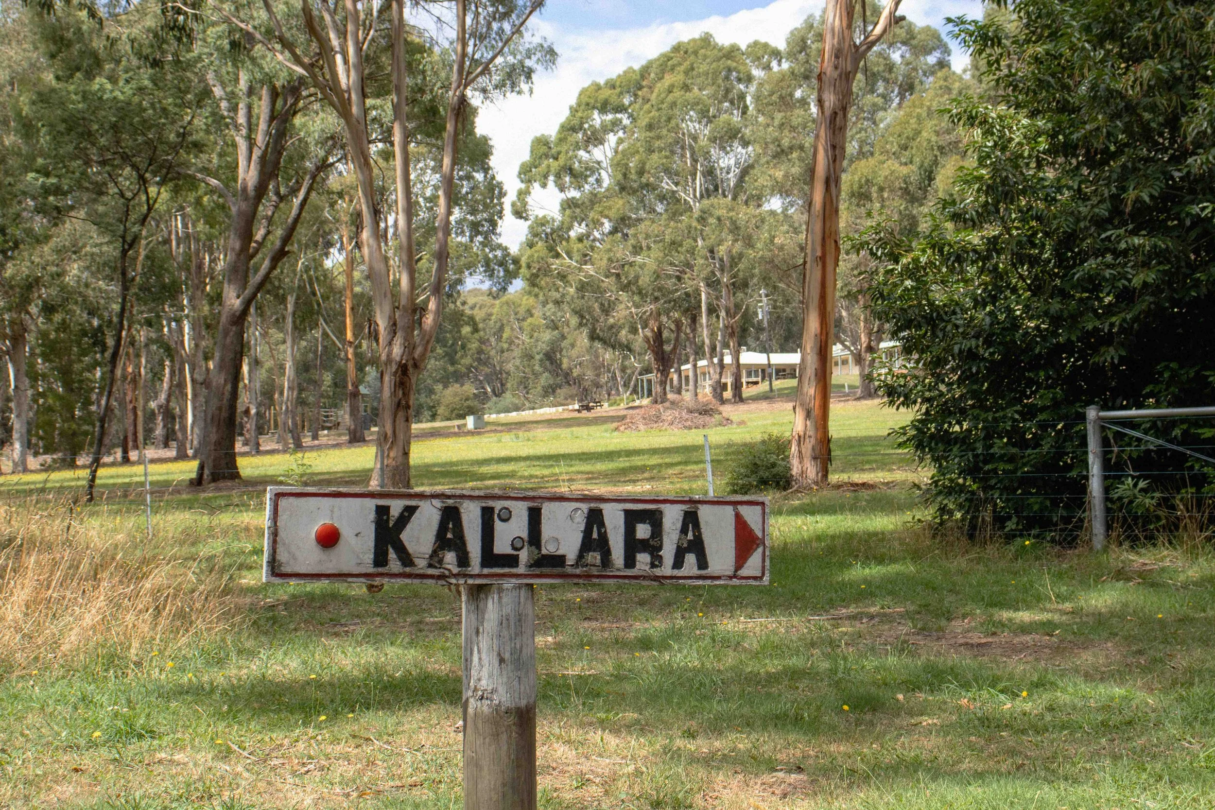 A farm or rural area with tall trees, a grassy area, and a white house in the background. In the foreground, there is a weathered sign that reads 'KALLARA' with a red dot and an arrow pointing to the right.