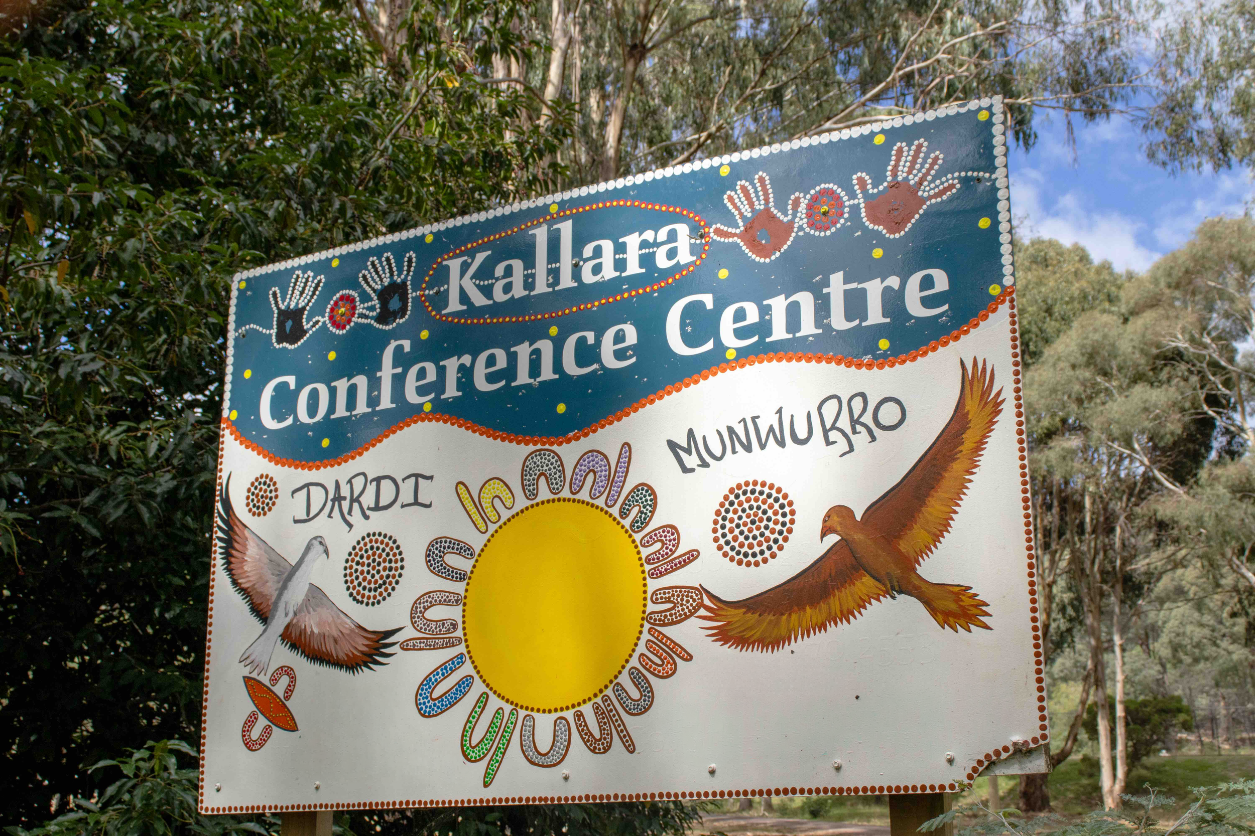 A decorative outdoor sign for the Kallarra Conference Centre, featuring Aboriginal art elements, birds, a sun, and surrounded by greenery and trees.