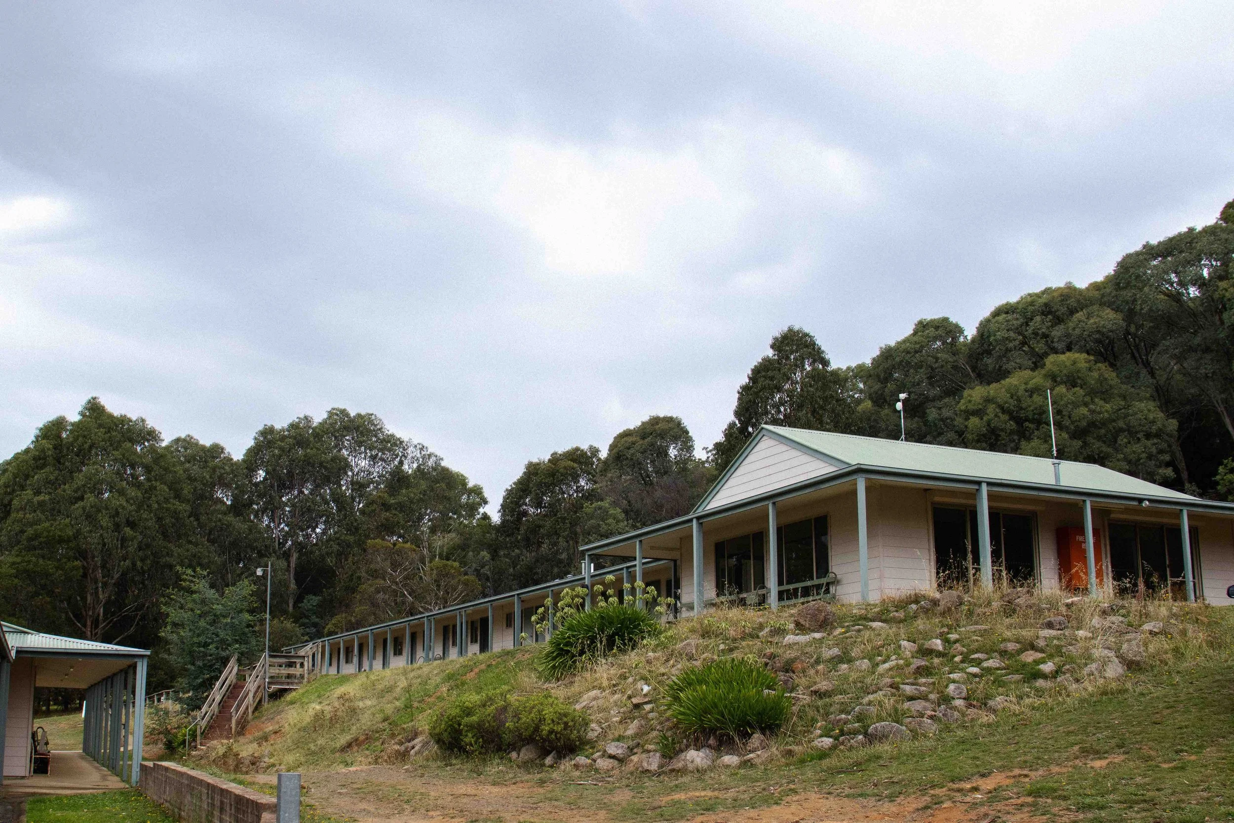 A building with a picnic shelter on a hillside with trees and cloudy sky in the background.