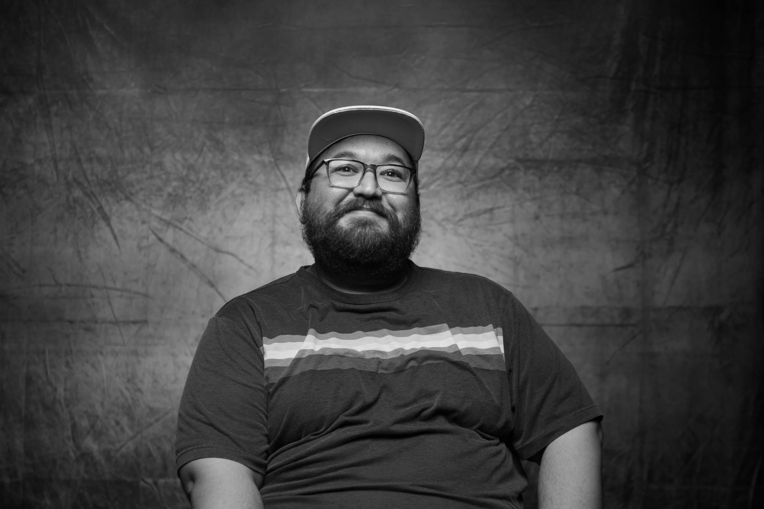 Black and white portrait of a man with glasses and a beard, wearing a cap and a t-shirt with horizontal stripes, sitting against a textured background.