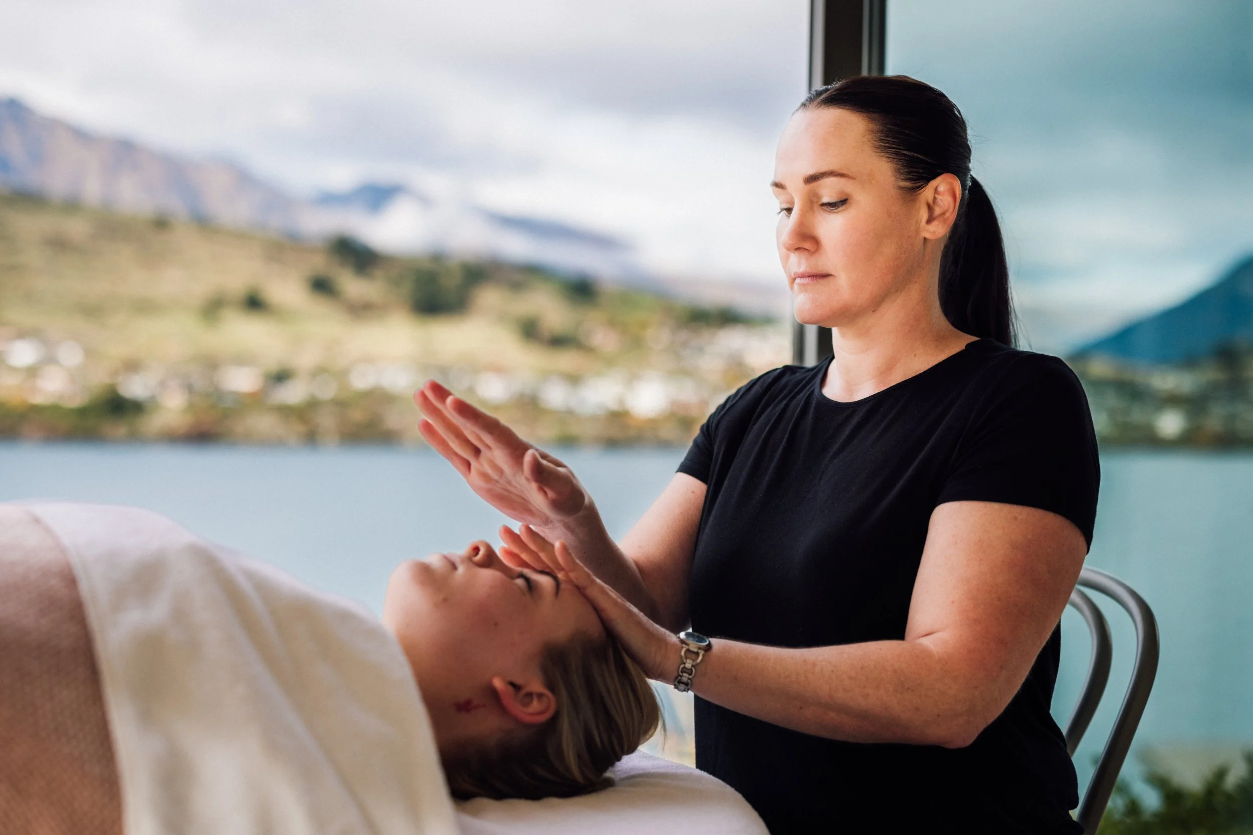 A woman with dark hair tied back, wearing a black shirt, providing a massage to a person lying down, in a room with large windows overlooking mountains and a body of water.