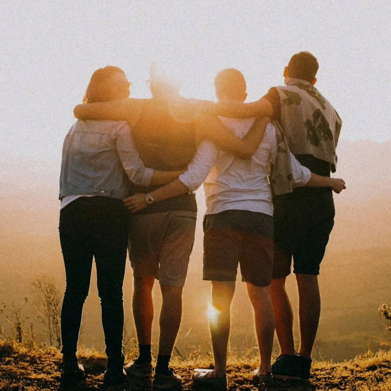 A group of four friends standing close together outdoors at sunset with arms around each other, viewed from behind.