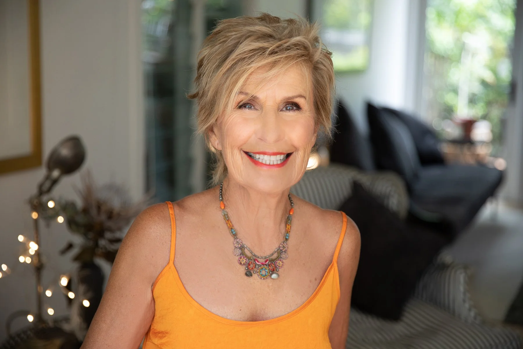 A smiling older woman with short blonde hair, wearing an orange spaghetti strap top and colorful beaded necklace, standing in a living room with natural light and modern furniture.