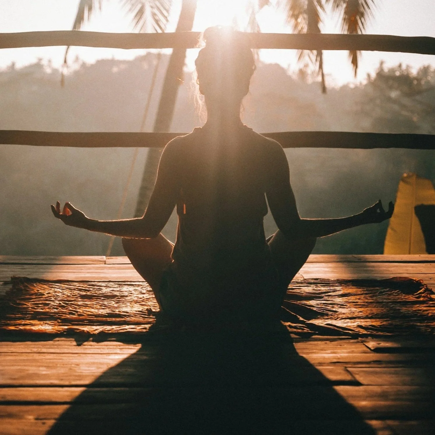 A person practicing yoga in a meditative pose at sunset on a wooden deck outdoors, with palm trees and a mountain in the background.