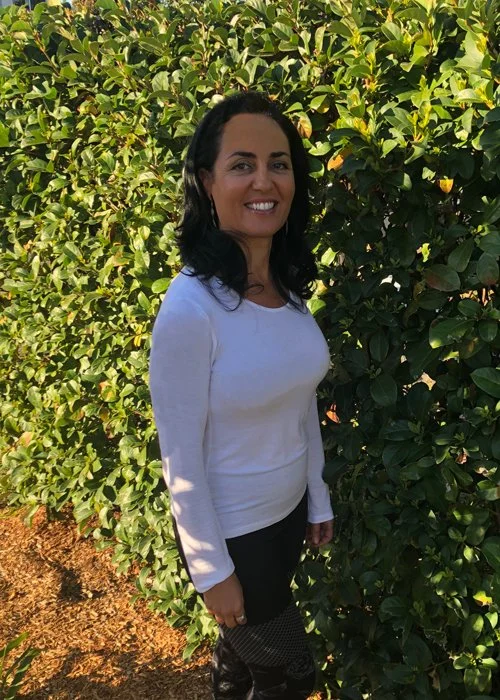 A woman standing outdoors in front of lush green bushes, smiling, wearing a white long-sleeve shirt and dark pants.