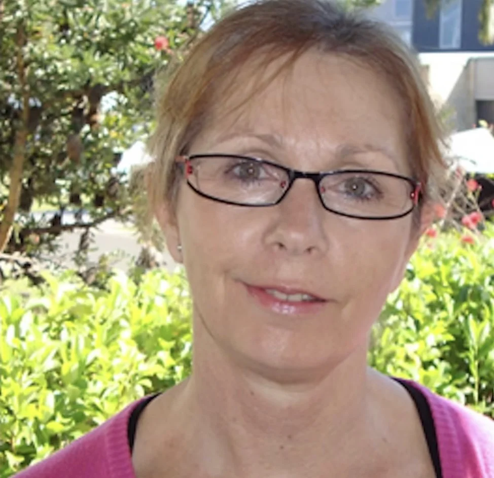 A woman with short reddish hair, glasses, and a pink sweater outdoors with greenery and a building in the background.