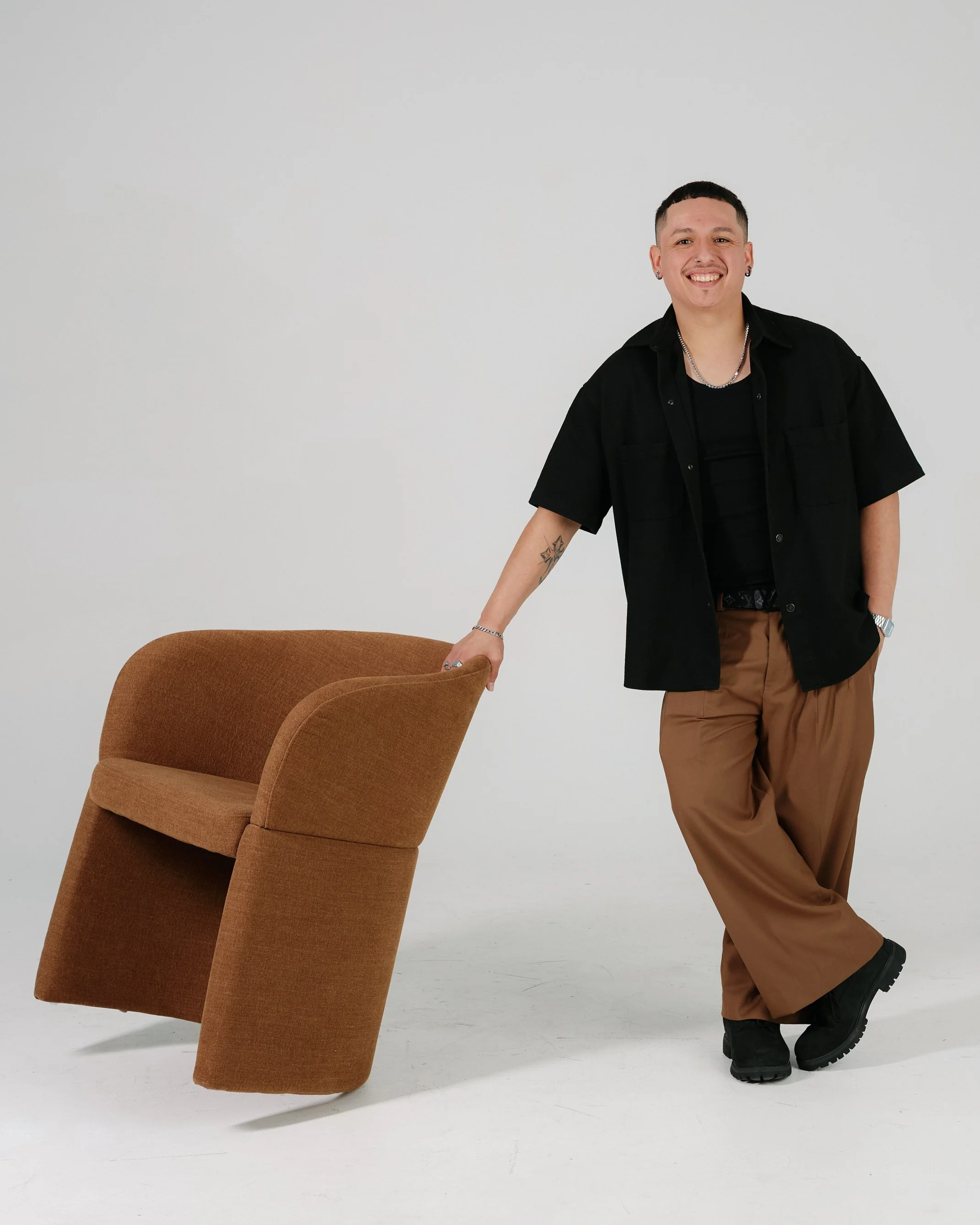 A man sitting on a high stool against a plain wall, wearing a dark jacket, turtleneck, and boots, smiling and looking to the left.
