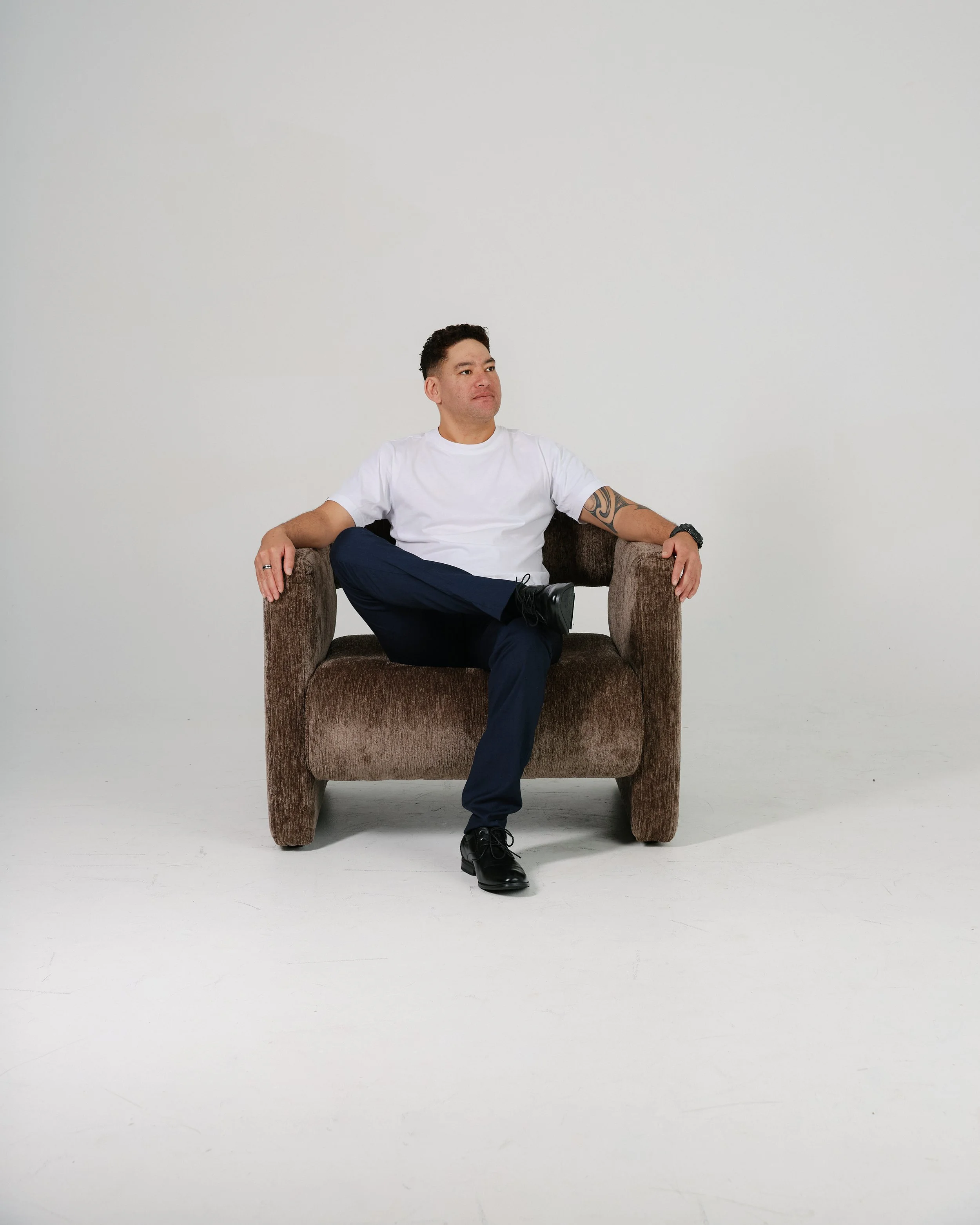 A man sitting in a brown armchair against a plain white wall, wearing a white t-shirt, dark pants, black shoes, and a tattoo on his left arm.