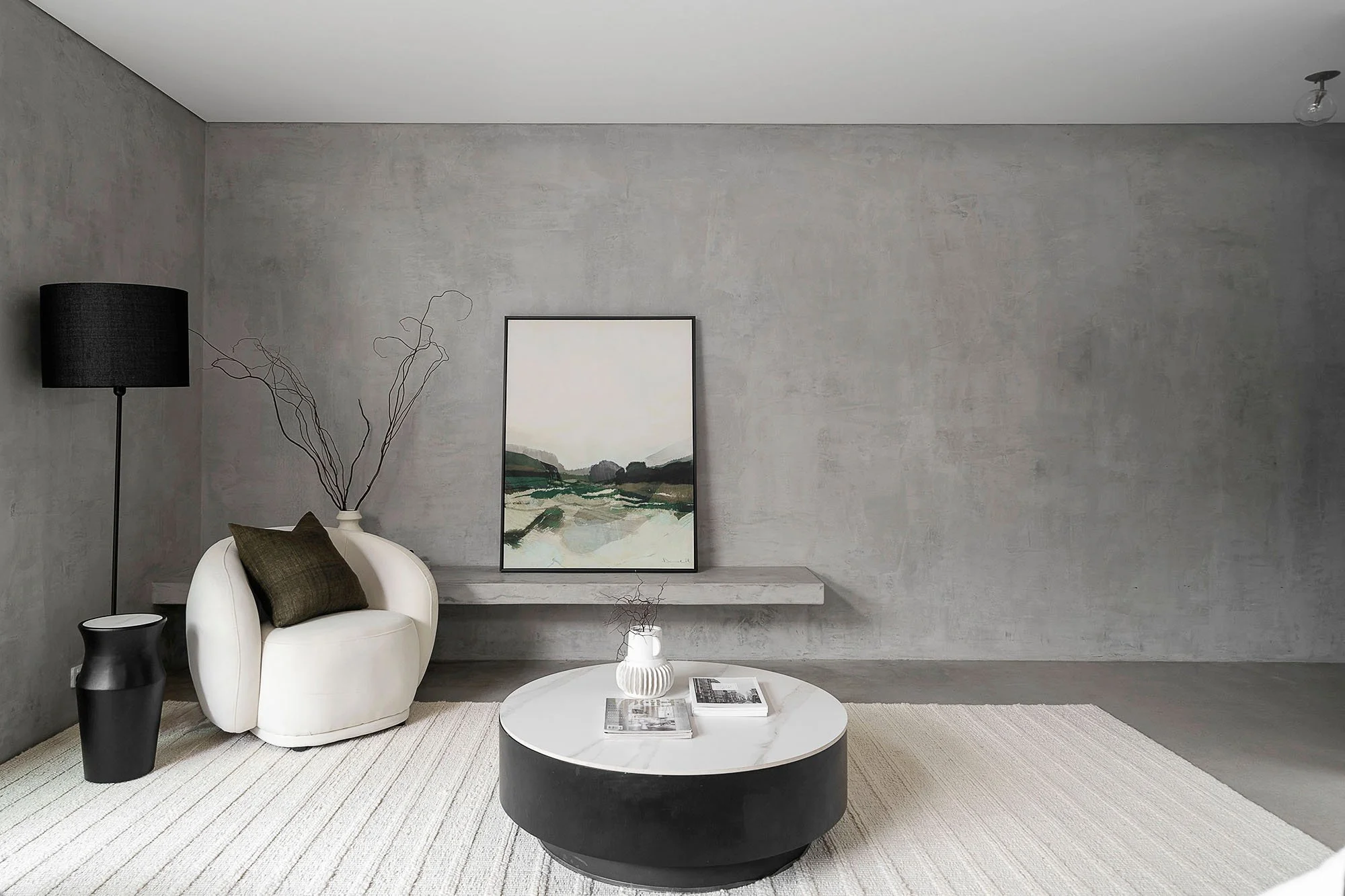Minimalist living room with a white armchair with a dark throw pillow, black floor lamp, abstract artwork on a gray wall, a round coffee table with books and a decorative vase, and a light-colored rug.