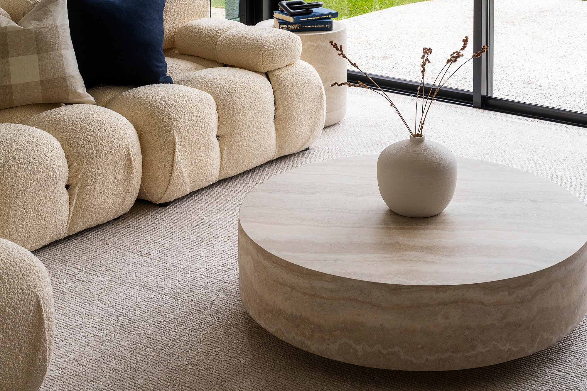 Living room with a textured cream sofa, plaid and dark cushions, a round marble coffee table with a white vase containing dried branches, and large glass sliding doors showing a gravel outdoor area.