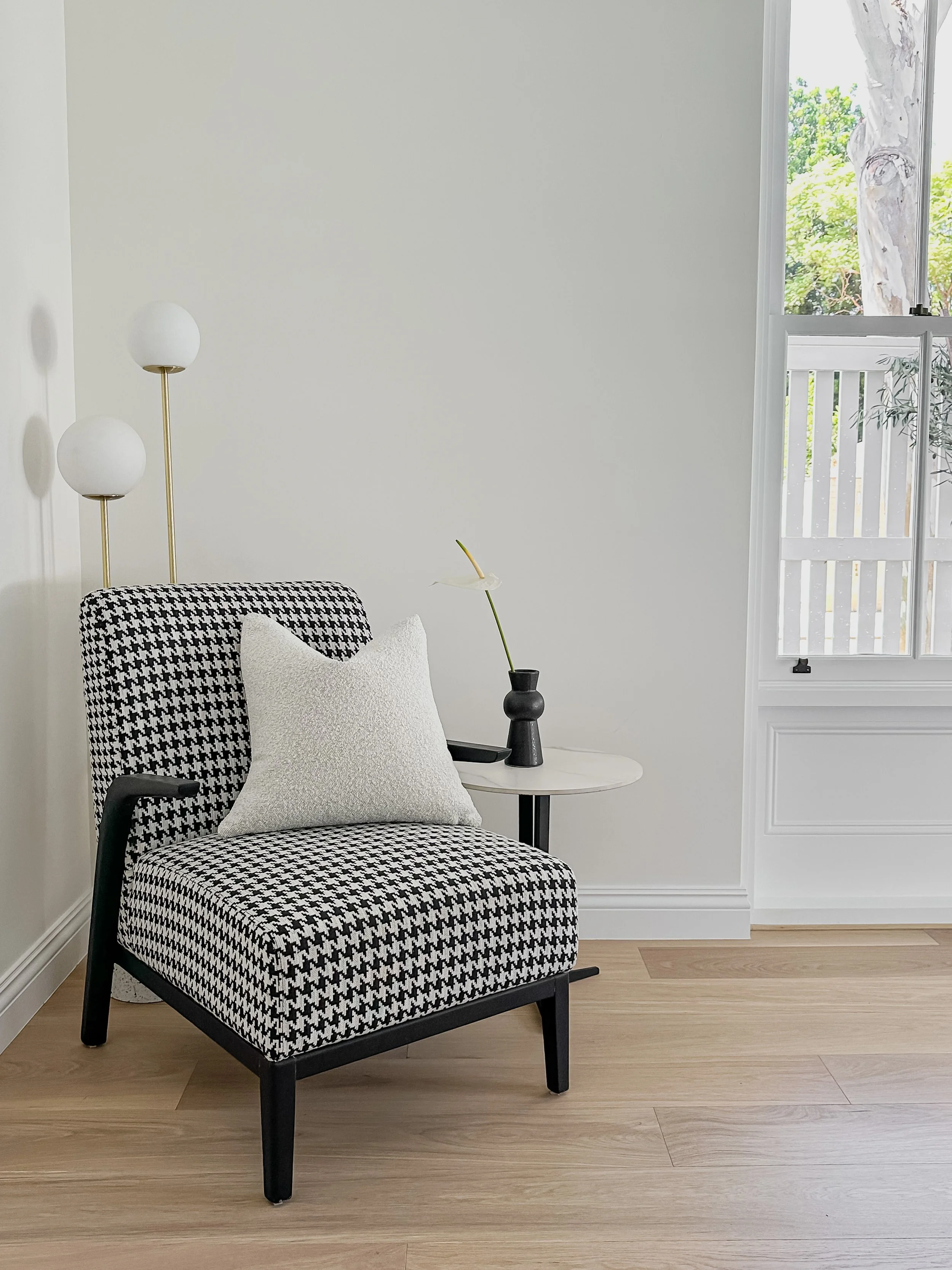 Black and white houndstooth-patterned armchair with a white textured pillow and a small white side table holding a black vase with a single long-stemmed flower, in a room with beige wooden flooring and a window with white trim and greenery outside.