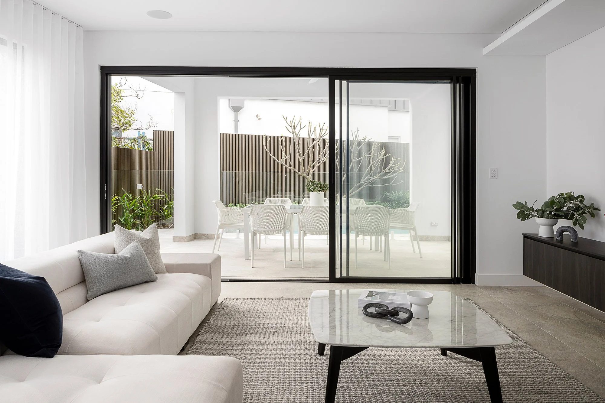 Modern living room with white sofa, coffee table, and sliding glass door leading to outdoor patio with dining set and plants.