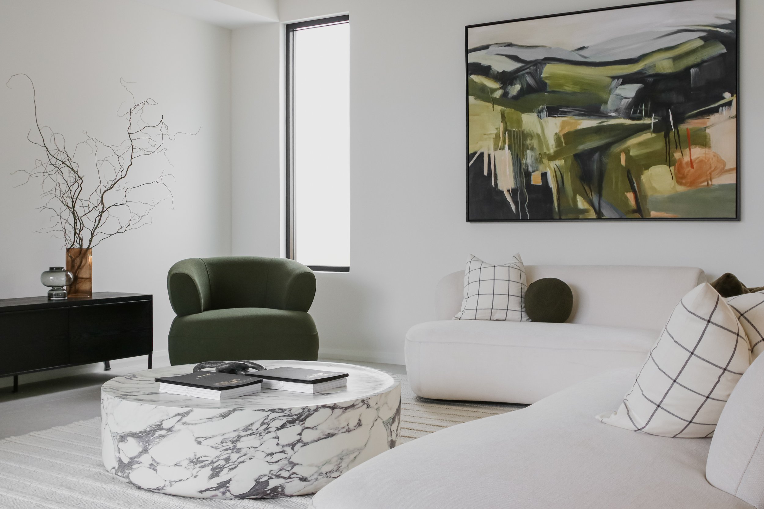 Modern living room with white walls, a white sofa with checkered and round pillows, a marble coffee table, a green armchair, abstract painting, and minimal decor.