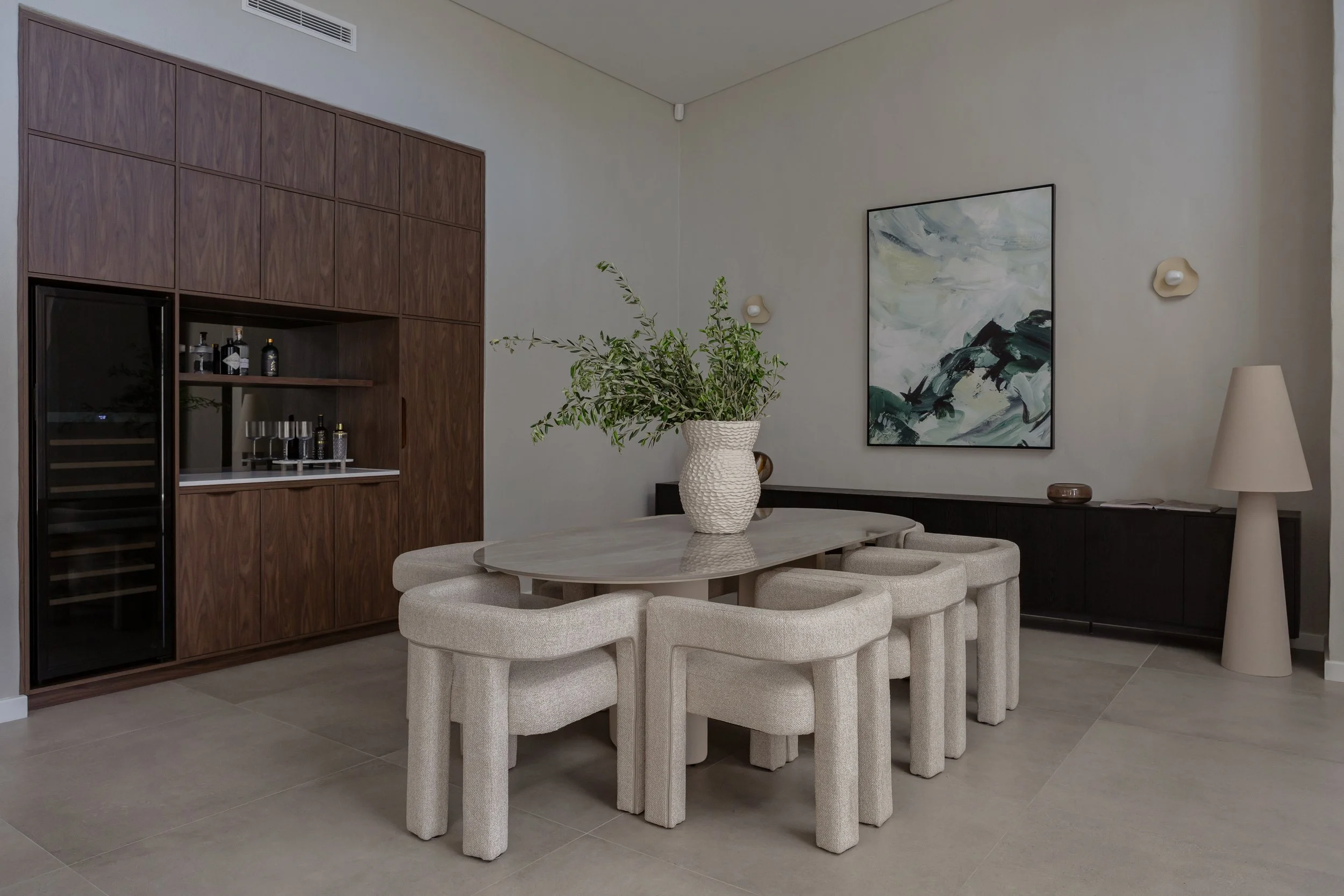 Modern dining room with a rectangular table surrounded by seven beige upholstered chairs. A large white textured vase with greenery is on the table. The back wall features a wooden cabinet with built-in shelves and a black wine cooler. A dark sideboa