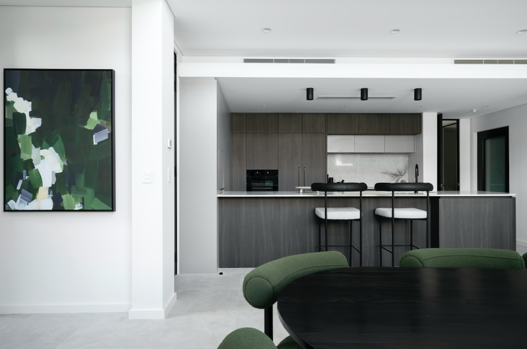 Modern kitchen with dark wood cabinets, white countertops, and two white barstools, adjacent to a dining area with green chairs.