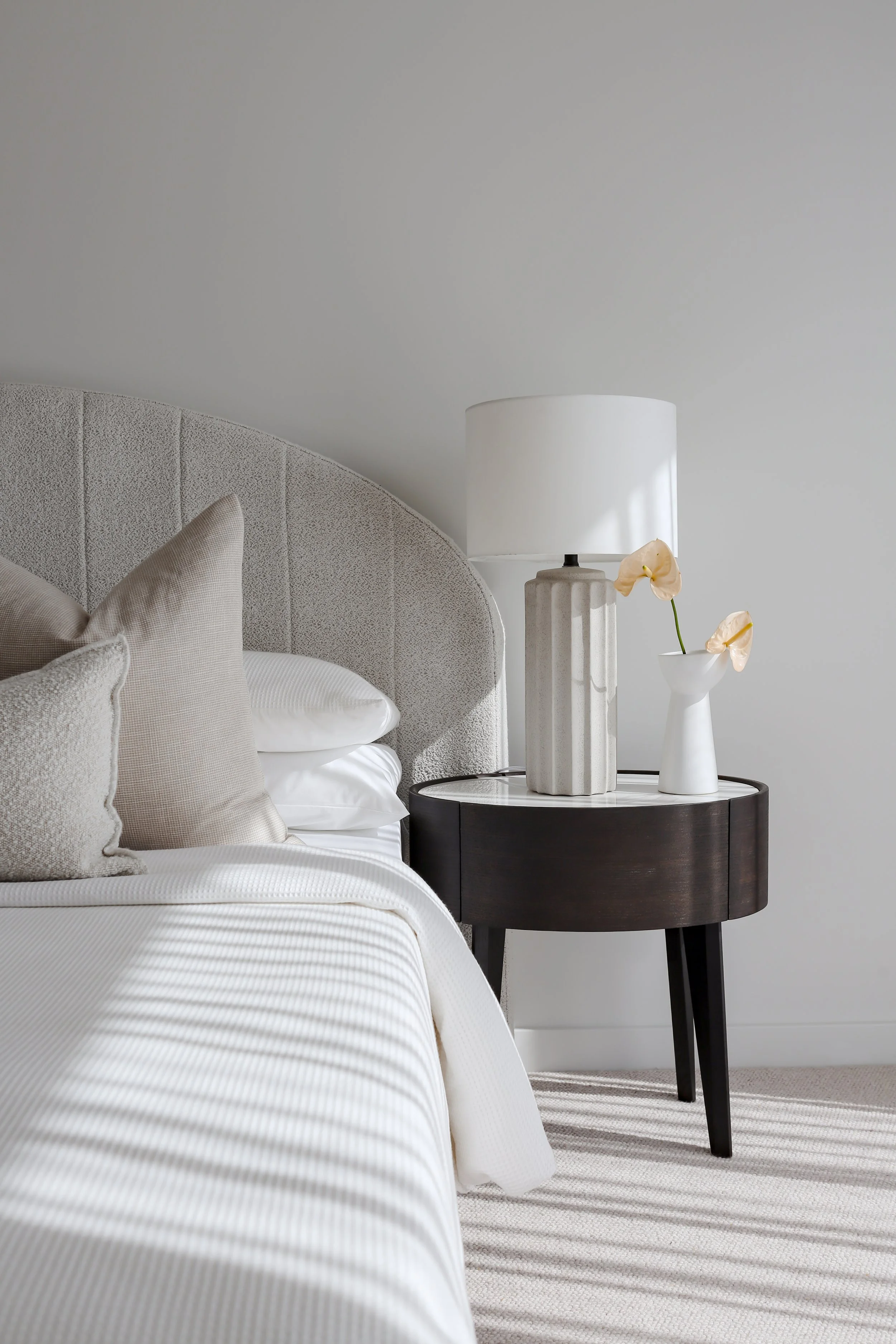A bedroom corner with a neutral-toned upholstered headboard, beige and white pillows, white bedding, a dark small round side table, a white table lamp, and a white vase with yellow flowers, all on a striped carpet.