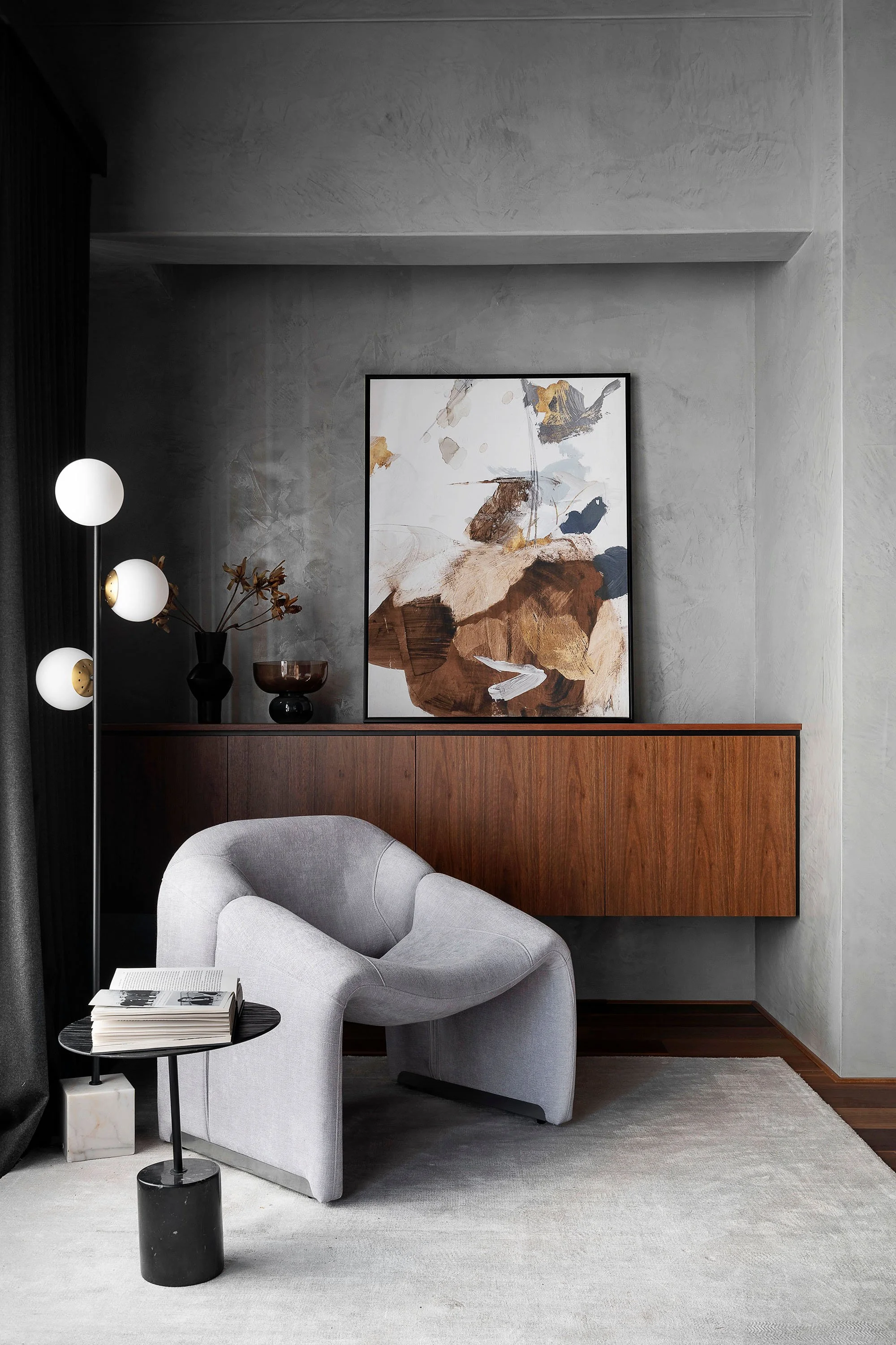 Modern living room corner with a light gray armchair, a small black side table holding a book, a wooden cabinet, abstract artwork on the wall, and decorative vases and a sculpture.