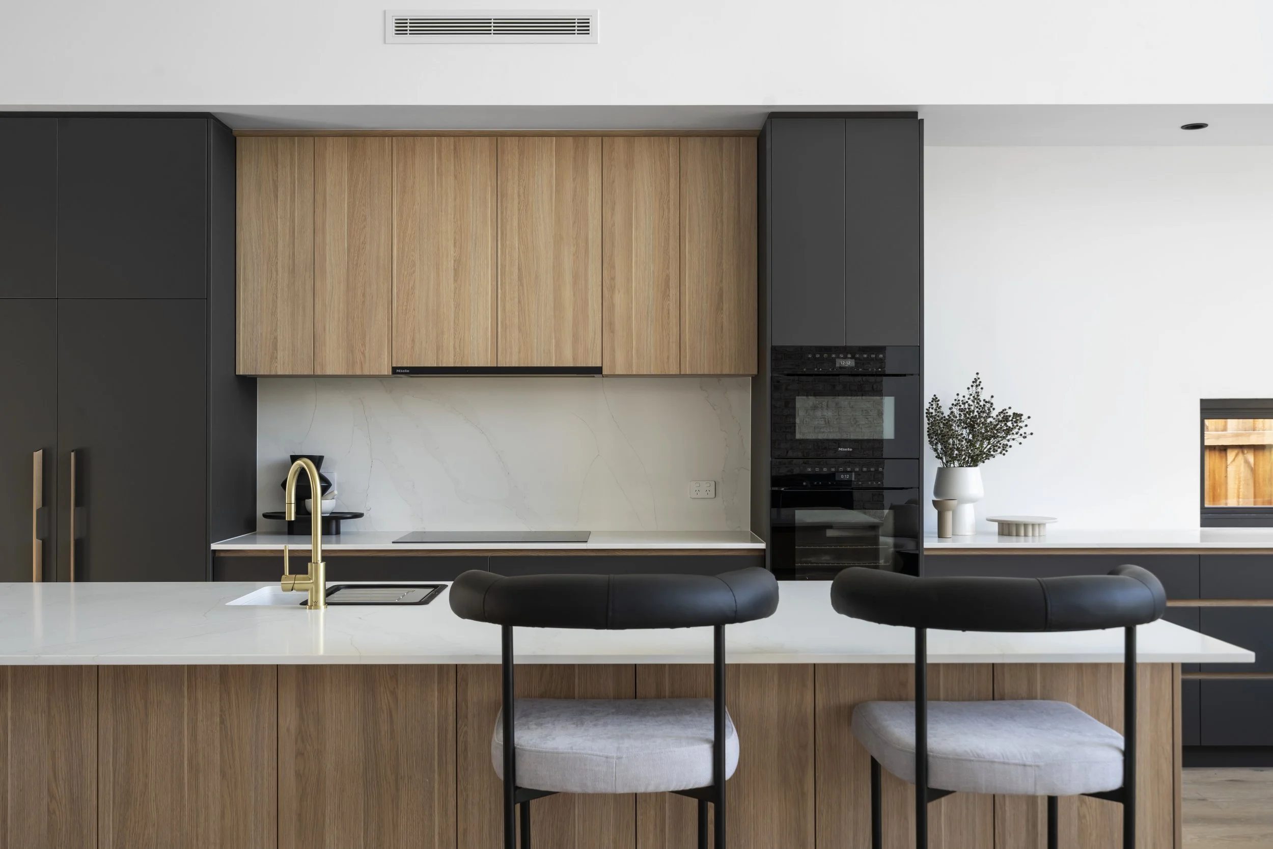 Modern kitchen with black and wood cabinets, white marble backsplash, gold faucet, black stools with white cushions, and a potted plant on the counter.