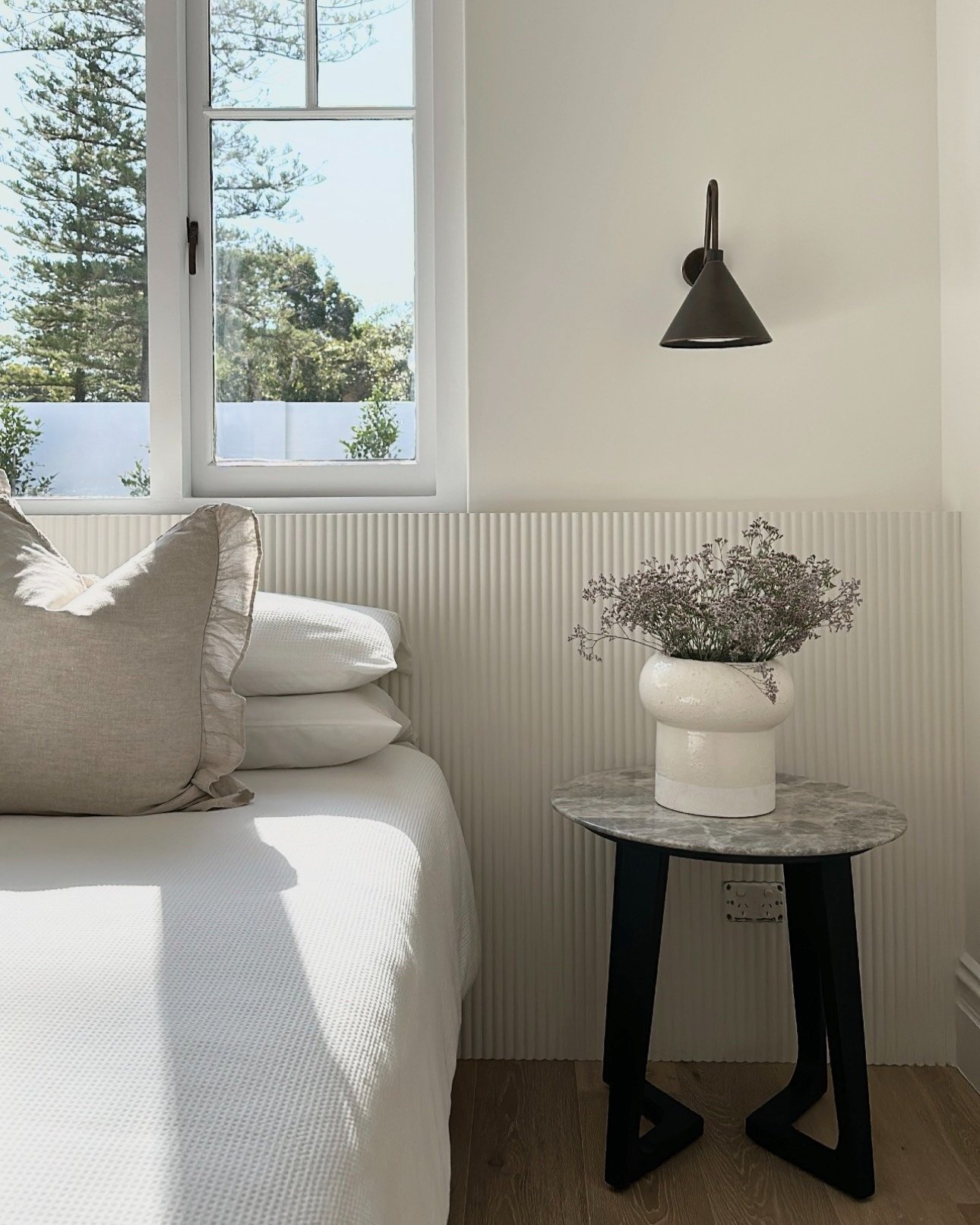 A white bed with beige pillows, a small round marble top side table with black legs, and a white vase of flowers on it, next to a window with trees outside, and a black wall-mounted reading lamp.