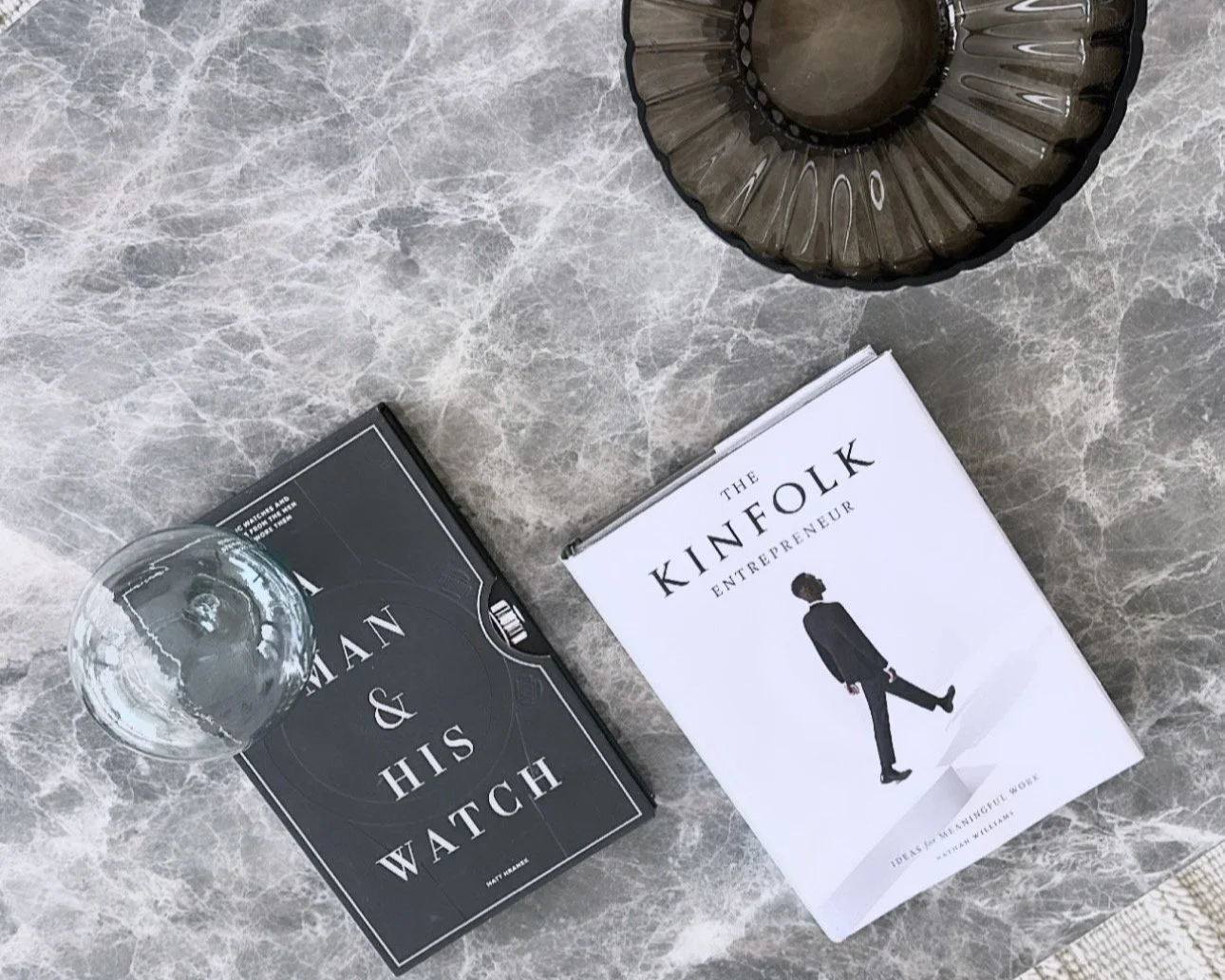 Books titled 'Man & His Watch' and 'The Kinfolk Entrepreneur' on a marble surface, with a glass of water and decorative bowls nearby.