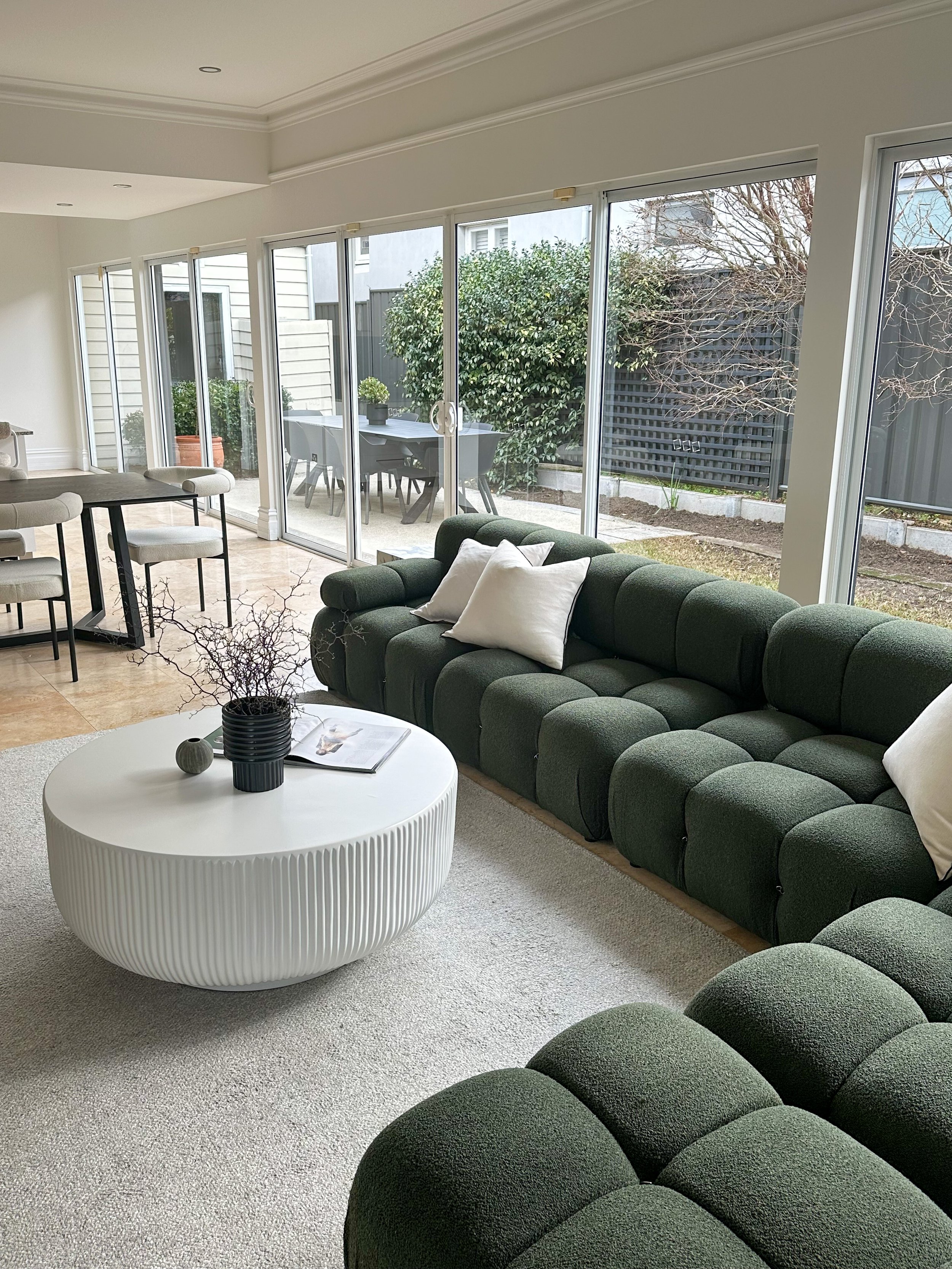 Living room with a green sectional sofa, white pillows, a white textured coffee table, and large glass sliding doors leading to a backyard patio.