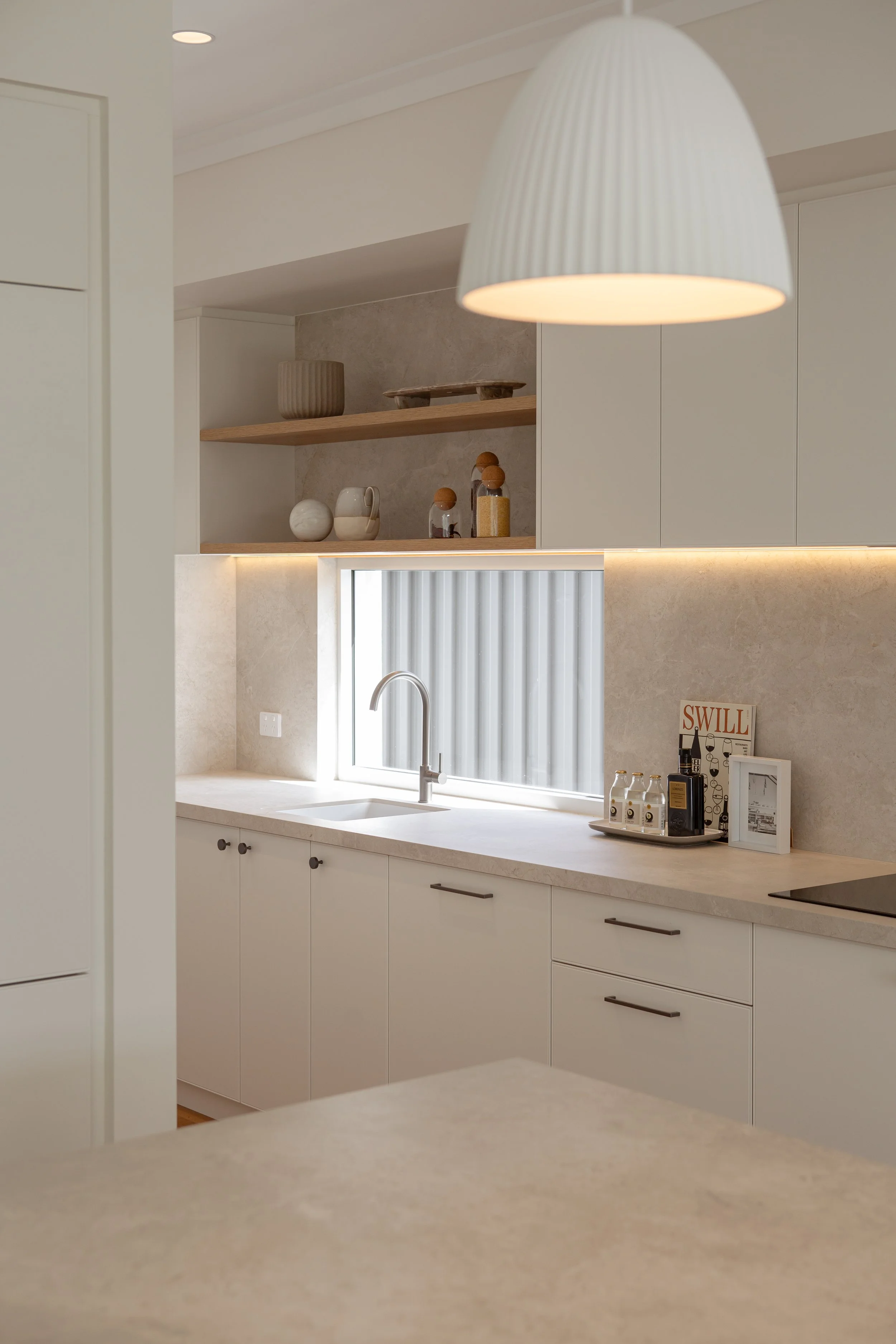 Modern kitchen with white cabinets, a countertop, and a window above the sink. A large white pendant light hangs overhead. There are decorative items and bottles on the counter and open shelves with vases and jars in the background.