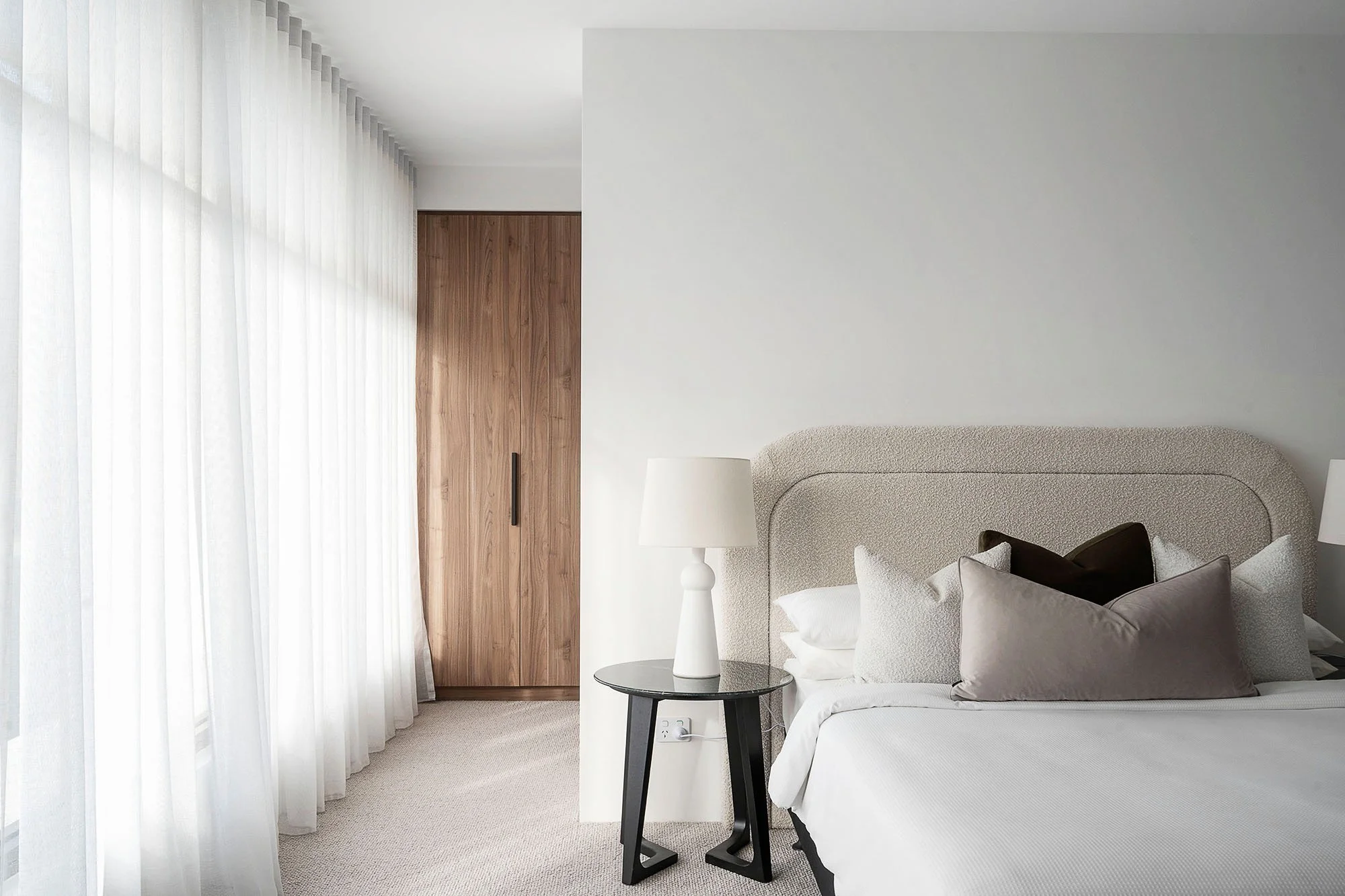 A neatly made bed with beige headboard, white bedding, and decorative pillows in black, gray, and beige. A black side table with a white lamp sits beside the bed. There are large white curtains covering windows and a wooden closet door in the backgro