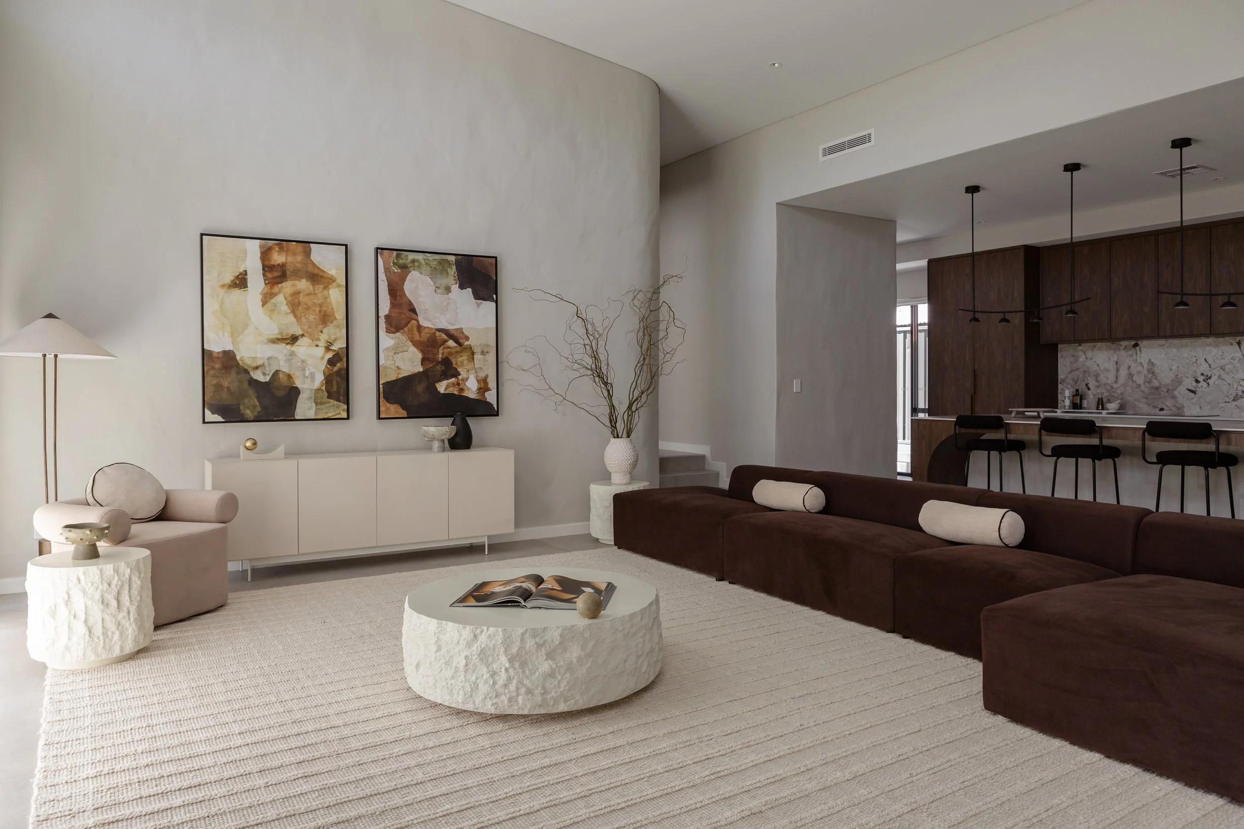 Modern living room with beige and brown furniture, abstract artwork on white wall, white textured rug, and kitchen area with bar stools in the background.