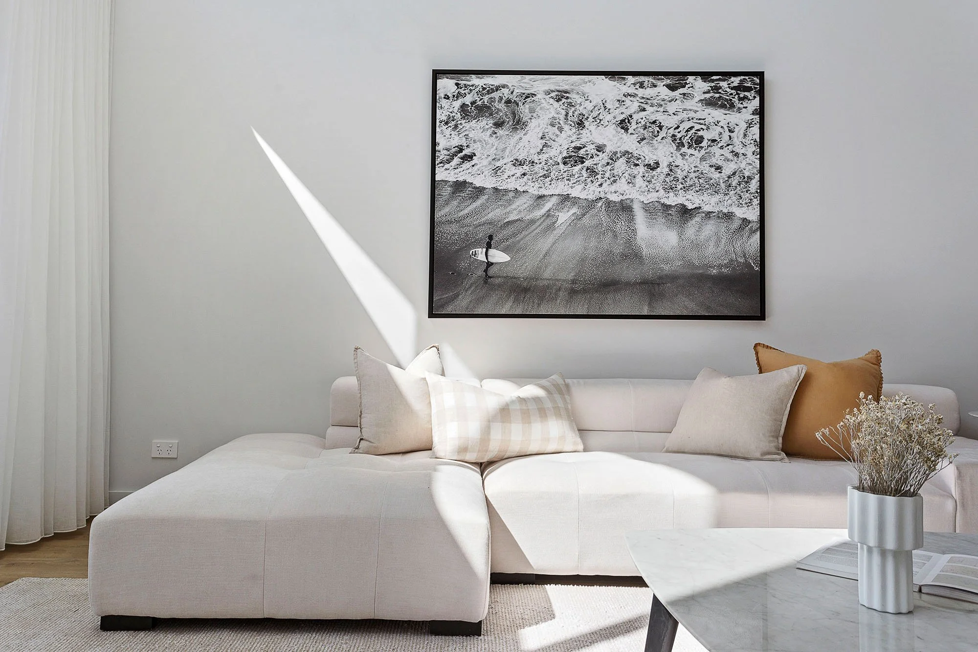 Modern living room with a white sectional sofa, beige and checkered pillows, a black and white ocean photo on the wall, a vase with white flowers, a marble coffee table, and sunlight streaming through a window.