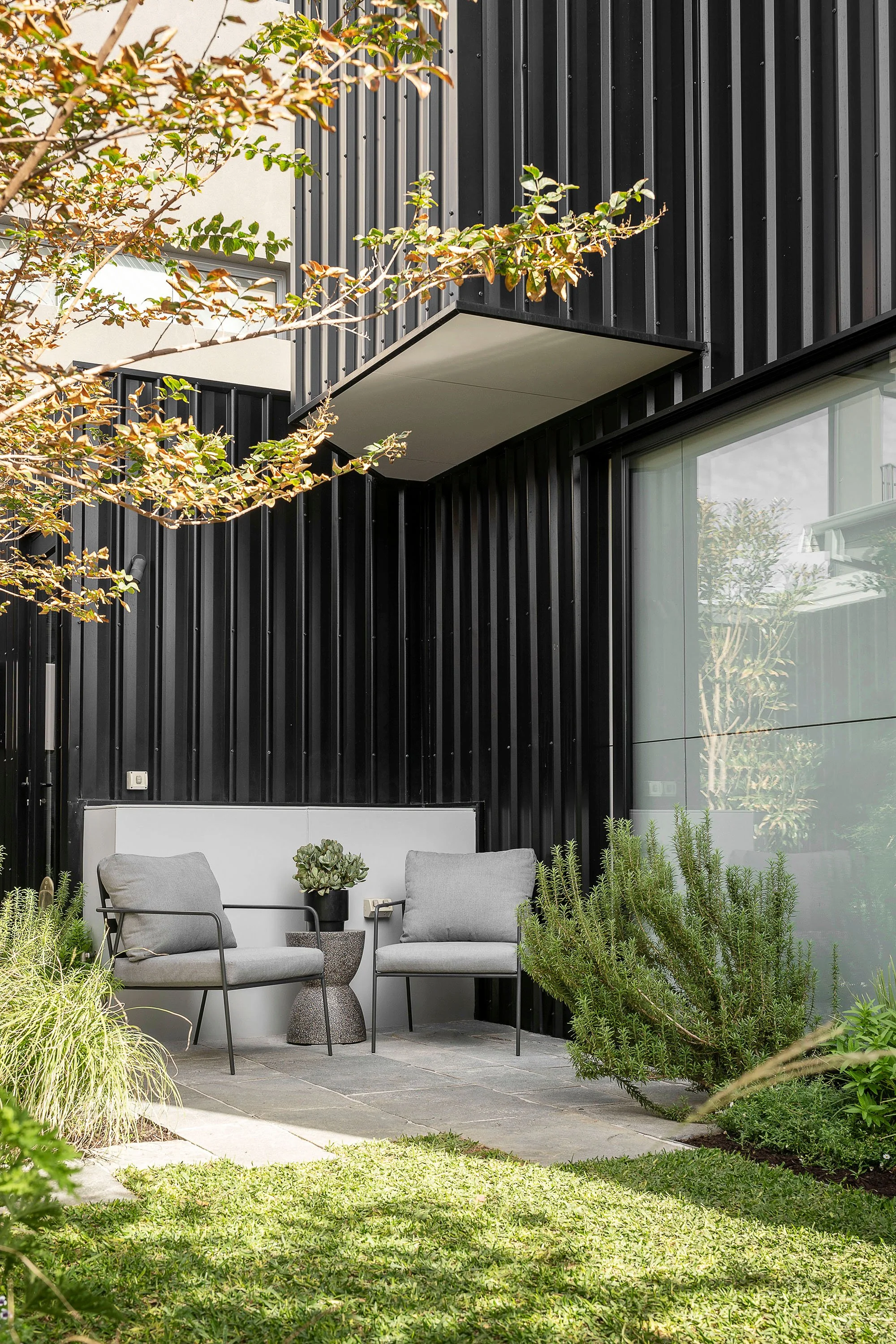 Modern black exterior wall of a building with a small patio area featuring two gray chairs, a small gray side table with a plant, and green shrubs and grass nearby.