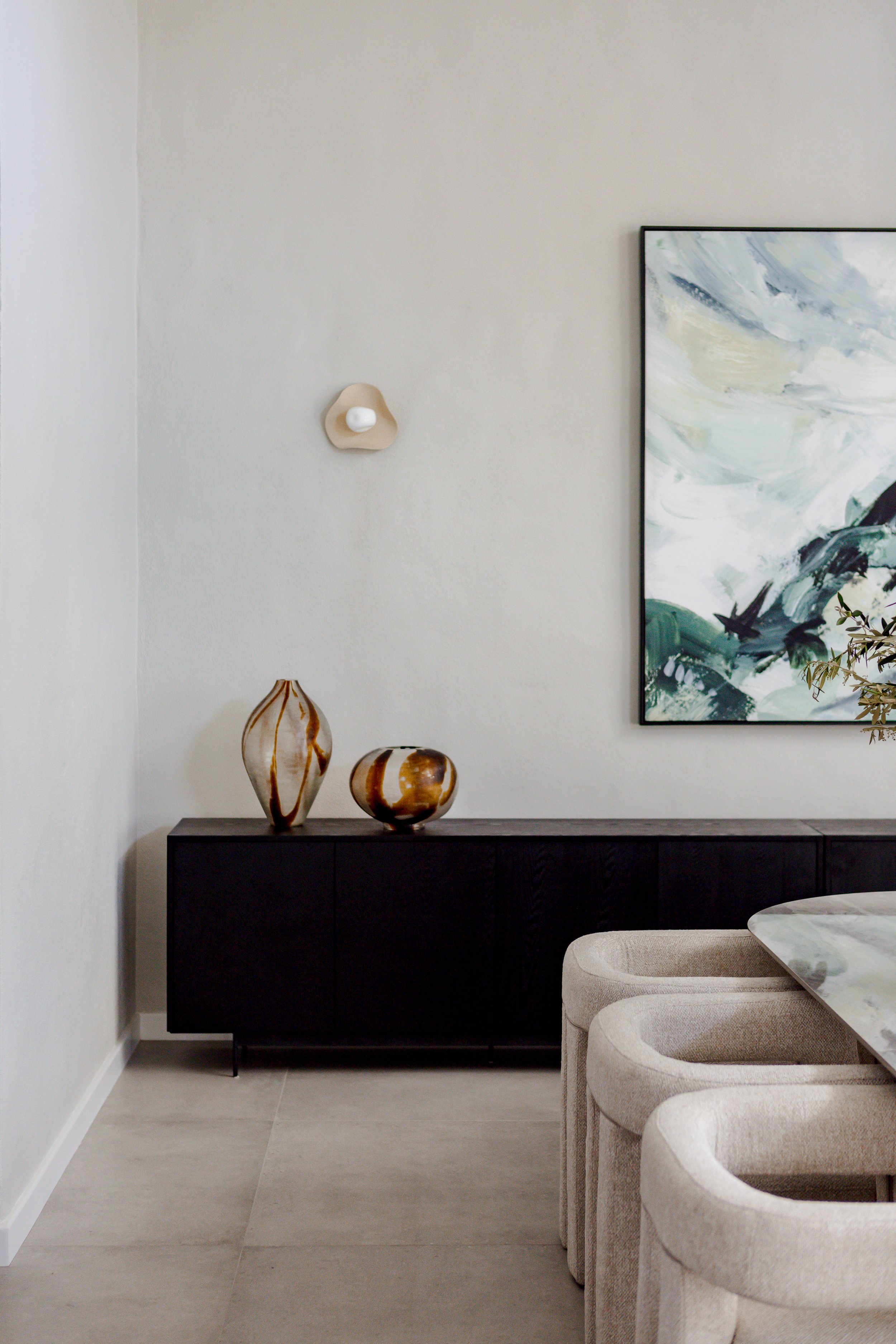 Interior of a modern living space with beige upholstered chairs, a black sideboard with two decorative vases, a large abstract wall art, and a wall-mounted lamp