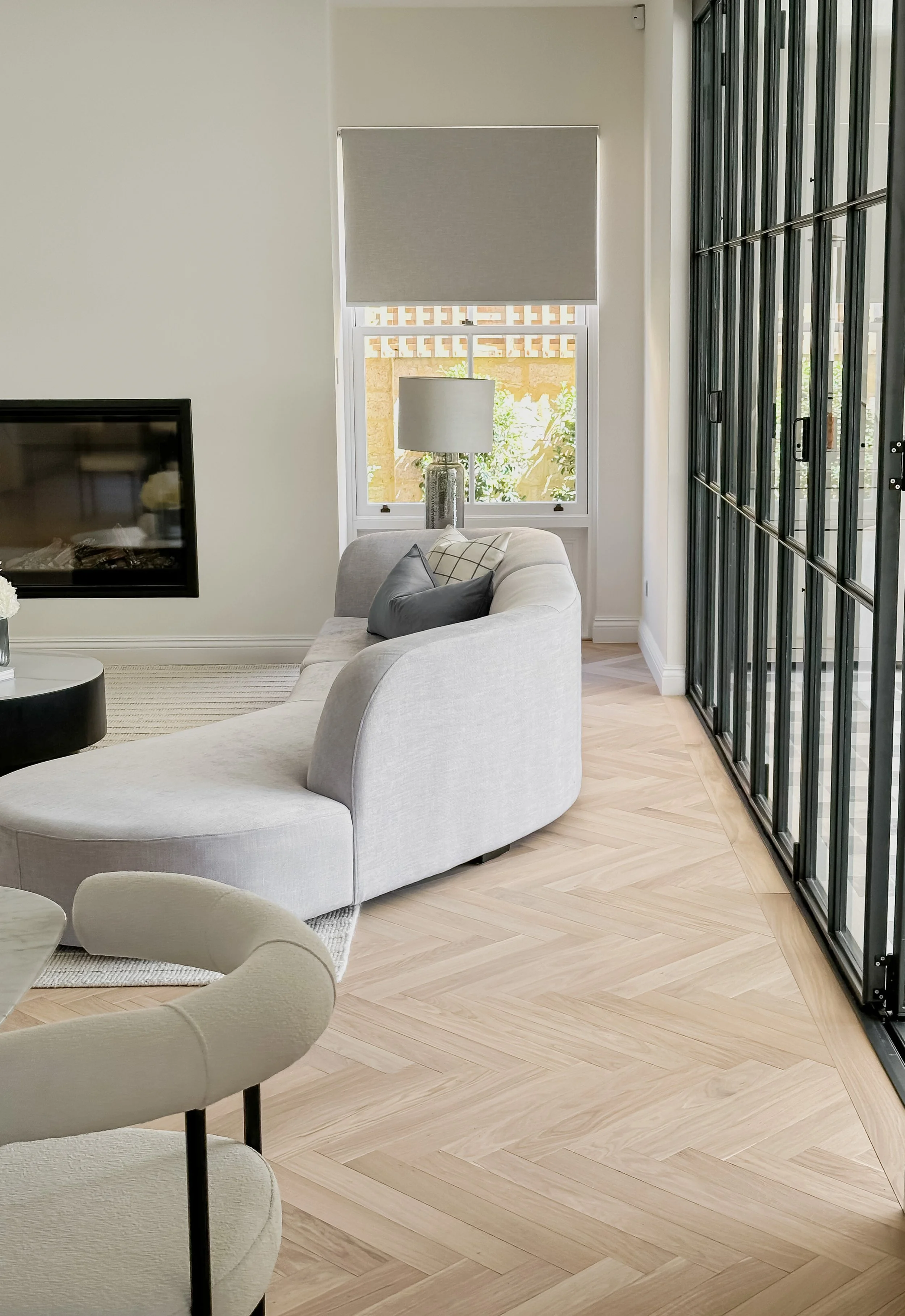 Modern living room with light gray curved sofa, black round coffee table, cream armchair, large window with blinds, and a glass sliding door.