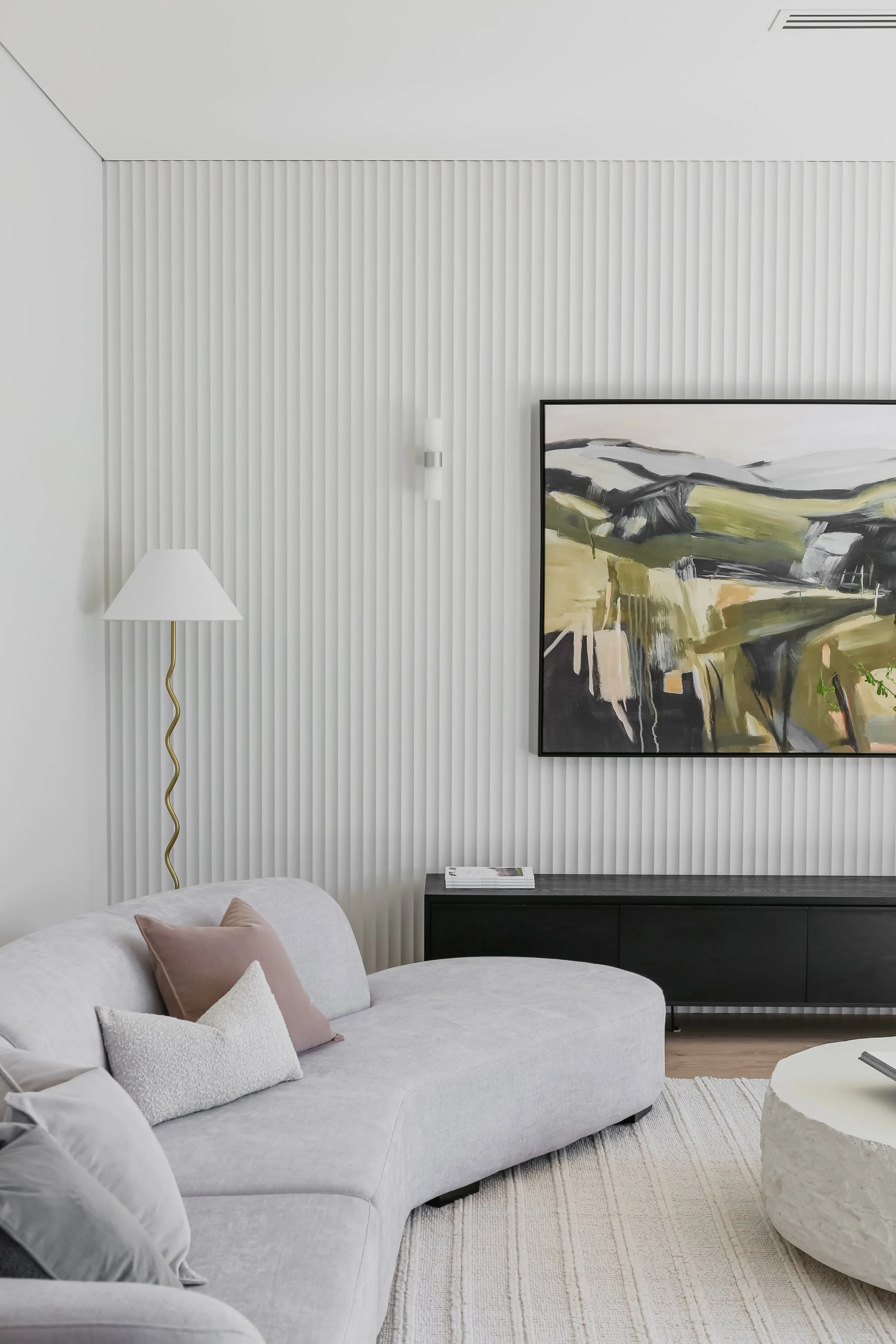 Modern living room with a white curved sofa and pastel throw pillows, black sleek media console, large abstract painting, striped white wall, and a tall floor lamp with a wavy gold pole in the corner.