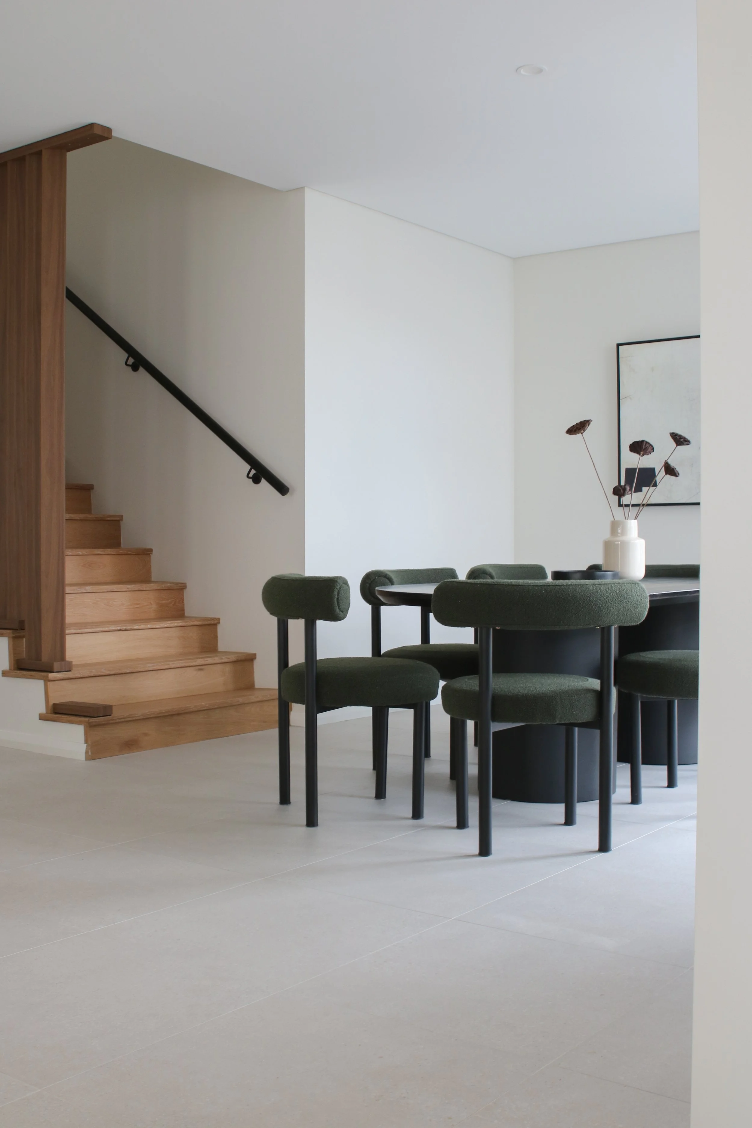 Minimalist dining area with a black round table surrounded by six green upholstered chairs, a white vase with dried flowers on the table, a large abstract art piece on the wall, light-colored tile flooring, and a staircase with wooden steps and black