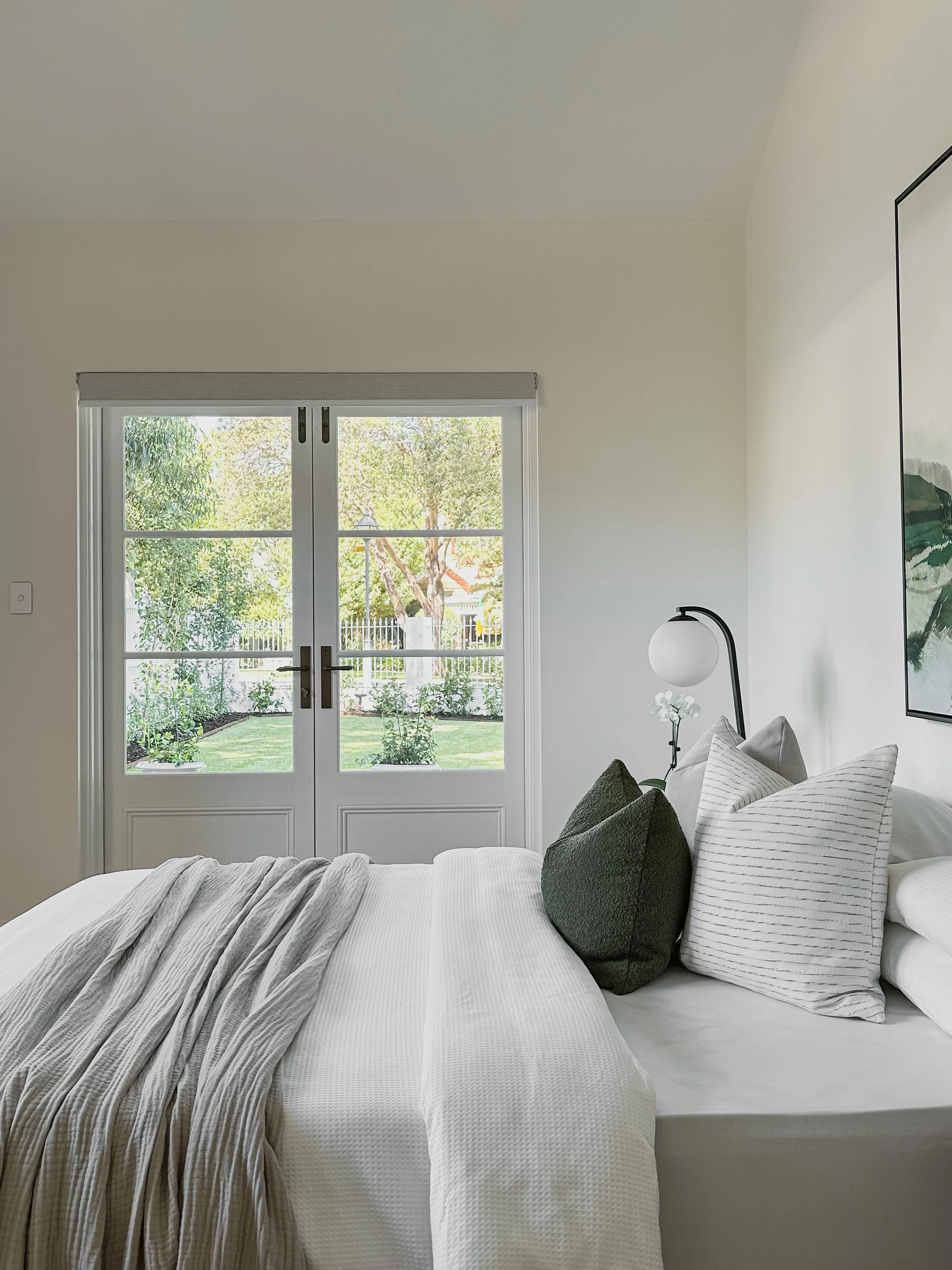A neatly made bed with white bedding and multiple pillows, next to a wall-mounted lamp and a large window showing a green garden outside.