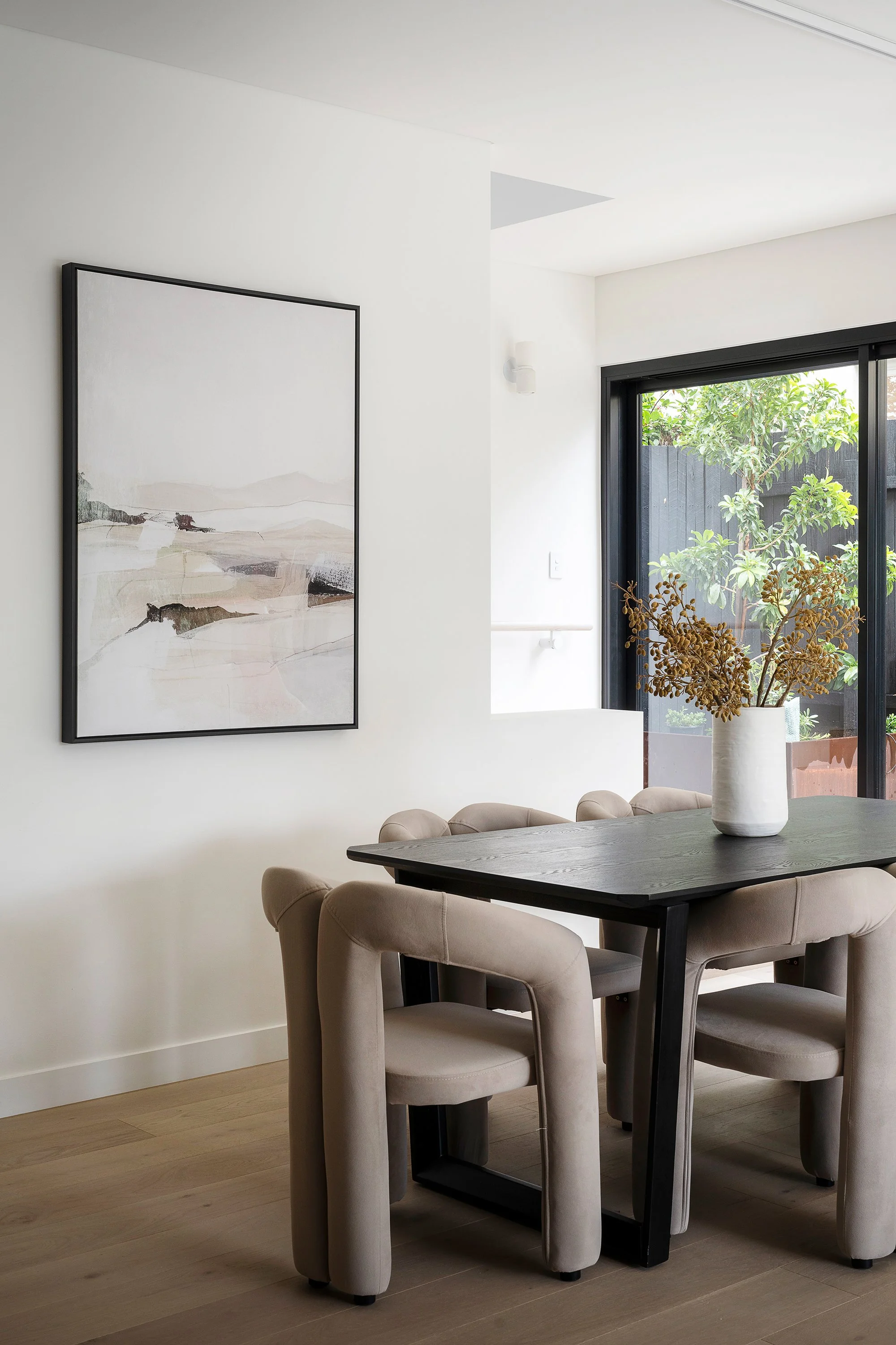 Dining room with a black table, beige upholstered chairs, a white vase with dried flowers, a large window with black framing, and a framed abstract painting on a white wall.