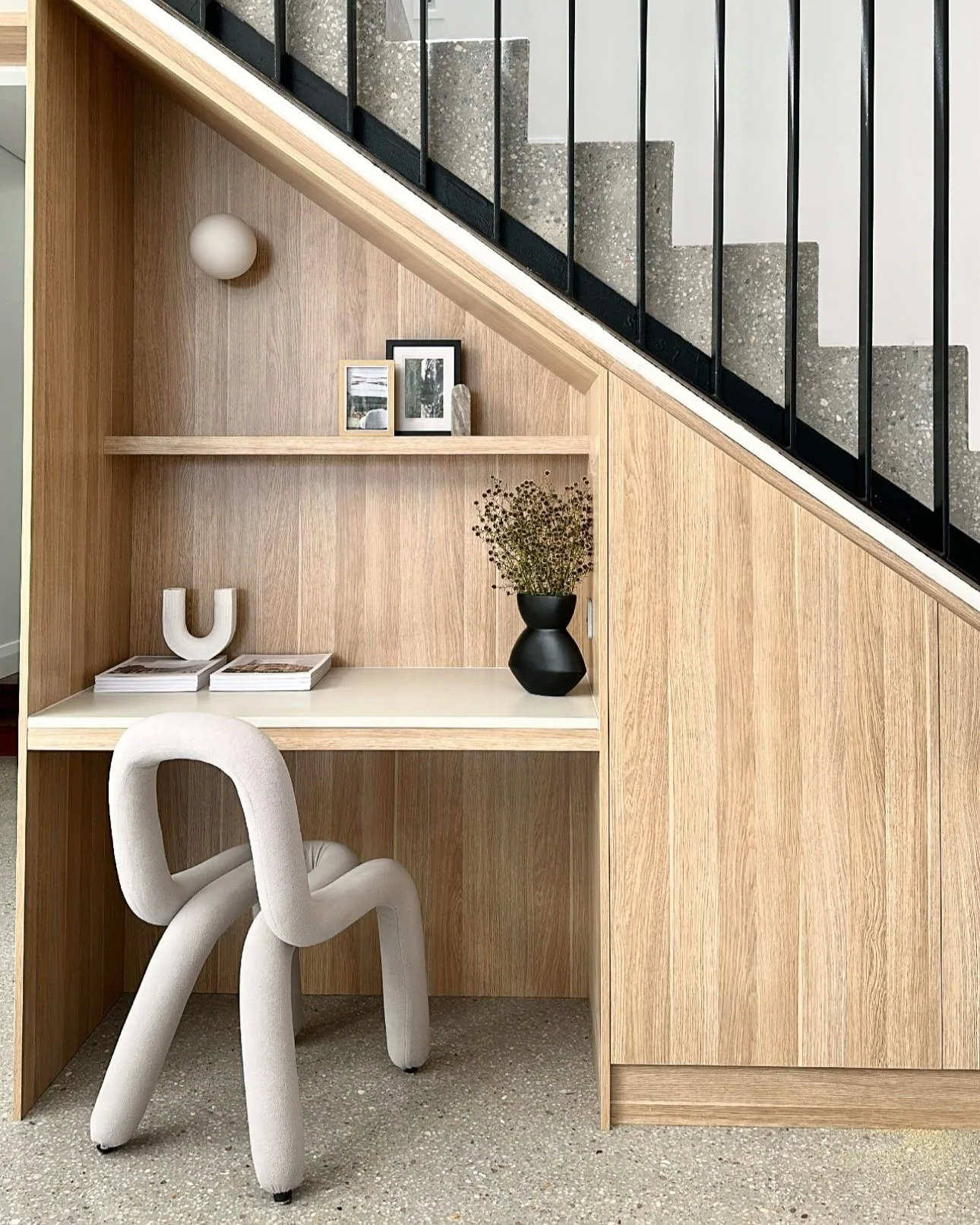 Corner of a room under a staircase with light wood built-in desk and shelves, a beige upholstered chair, decorative items including a black vase with dried flowers, framed photographs, a white ceramic 'U' sculpture, and a spherical wall light.