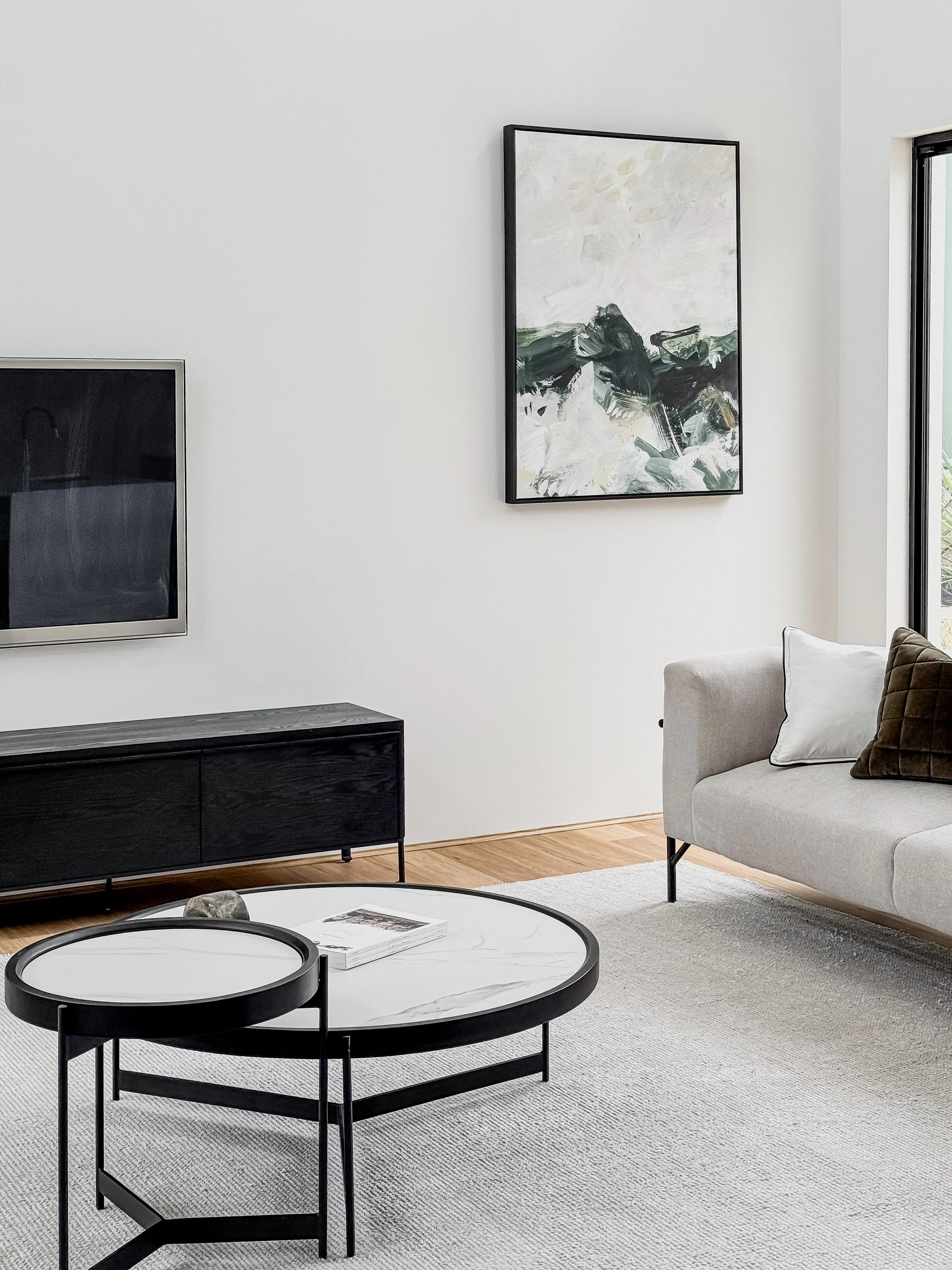 Modern living room with a gray sofa, abstract painting on white wall, wall-mounted TV, black and white coffee table, and wooden floor.