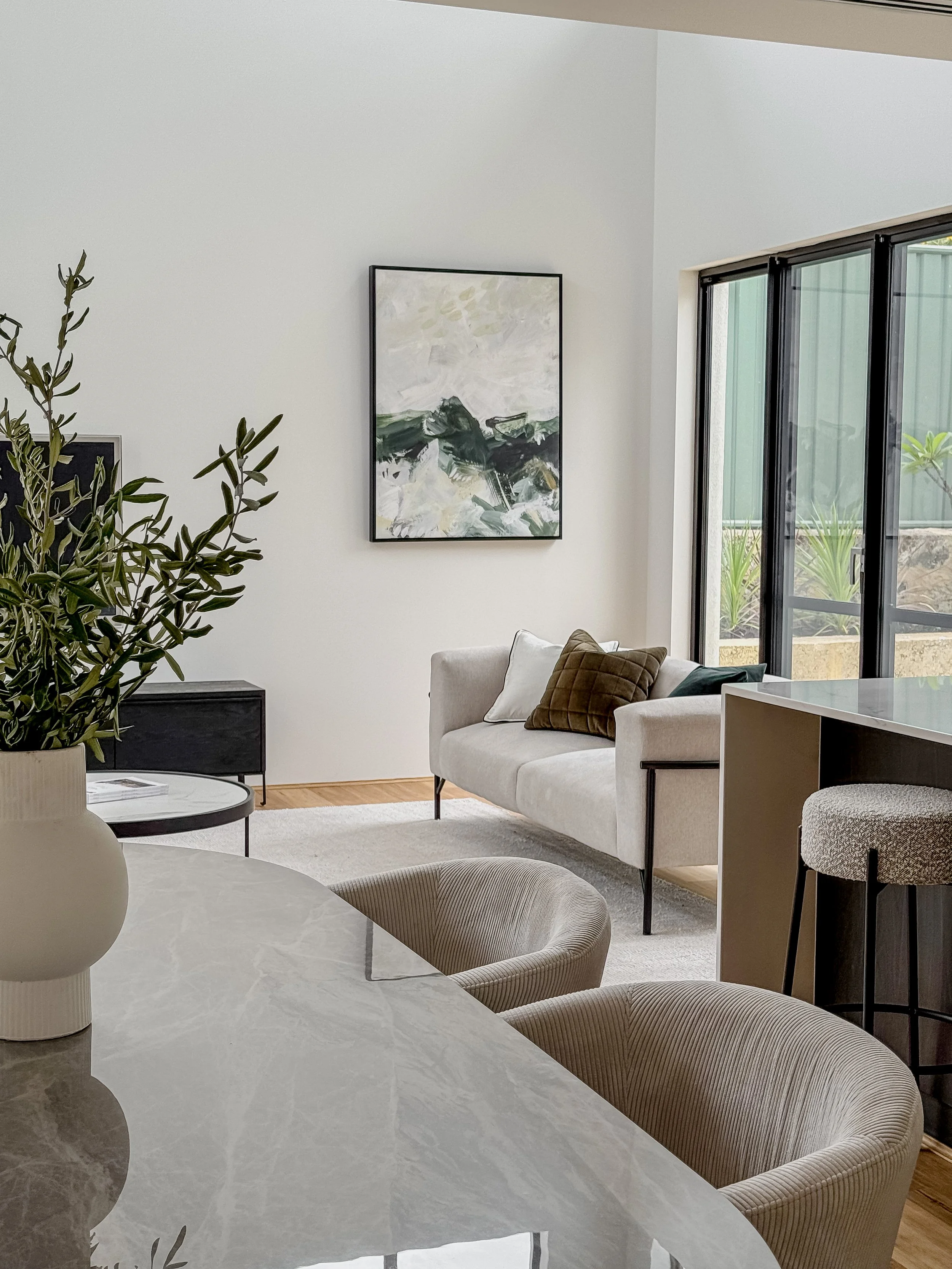 Modern living room with beige sofa, abstract artwork on white wall, glass sliding doors, and dining table with beige chairs and a decorative vase.