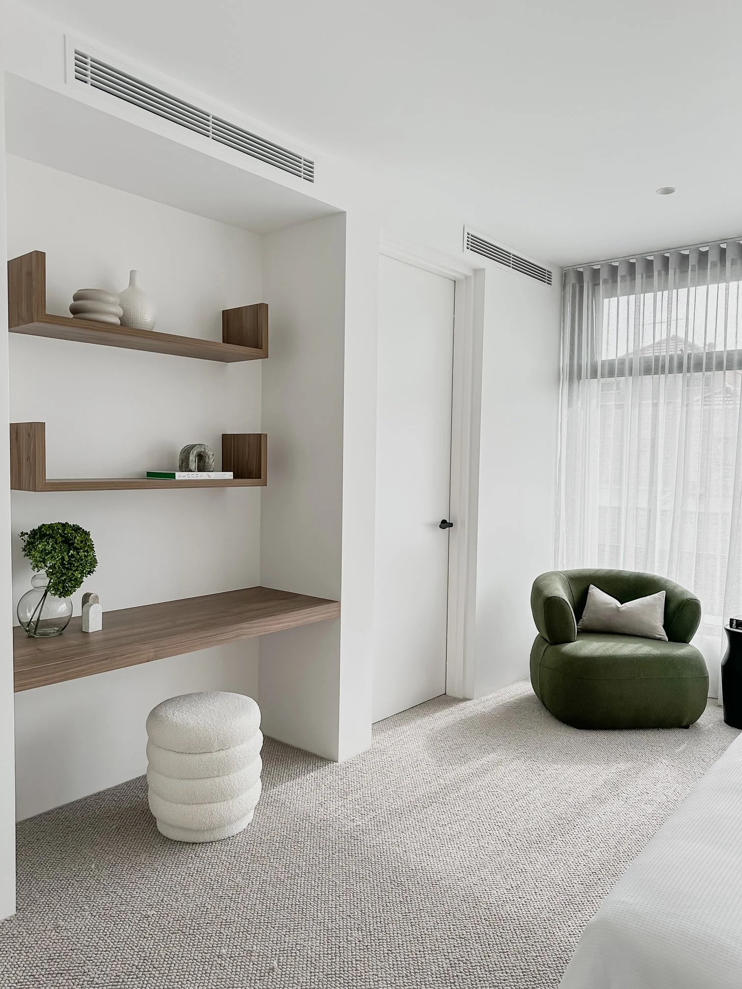Modern hotel room with white walls, beige carpet, a green armchair with a white pillow, sheer curtains, a built-in desk with a white cushion stool, and wooden shelves with decorative items.