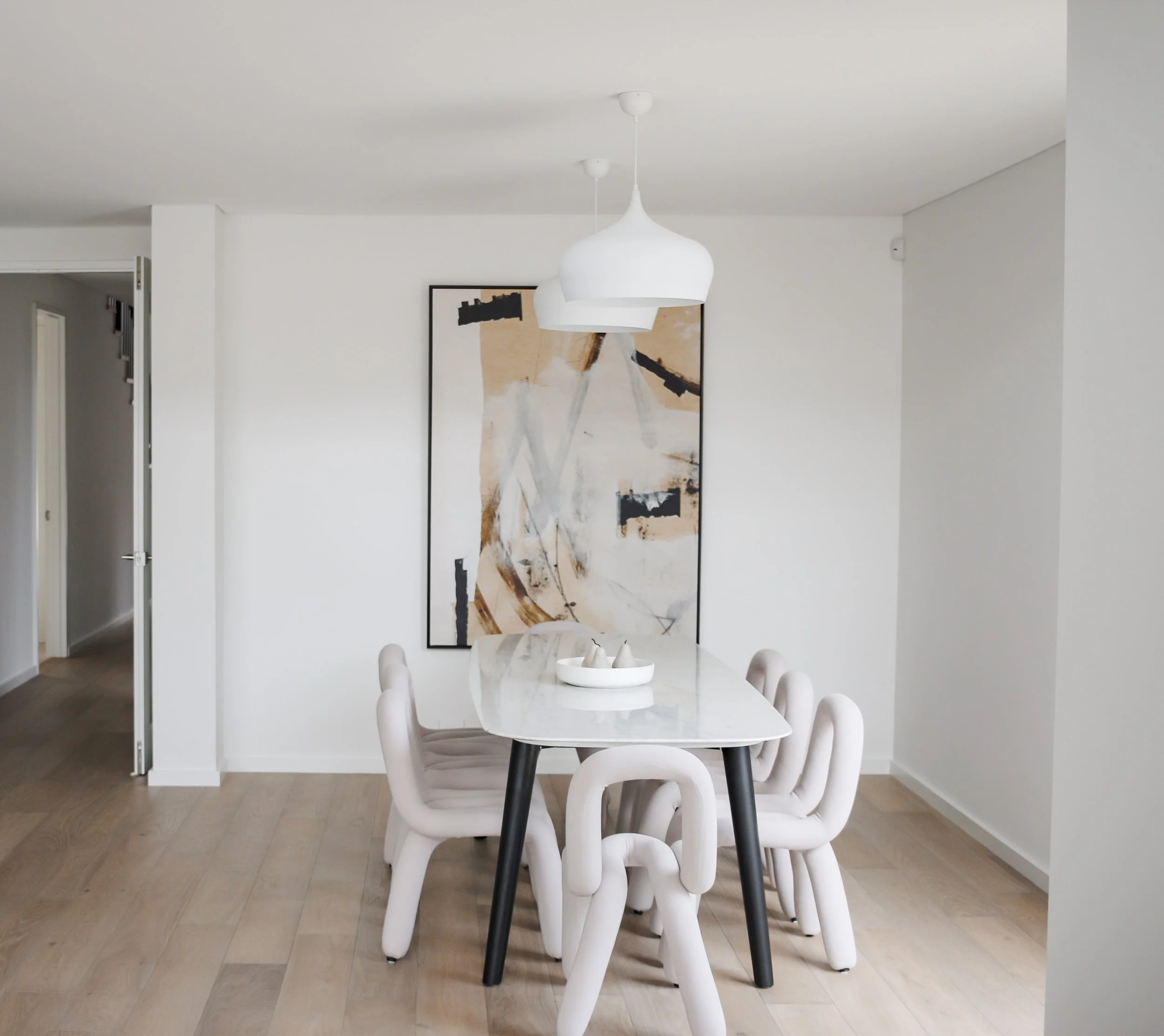 Modern dining room with a marble table, six unique white chairs, abstract wall art, and hanging white pendant lights.