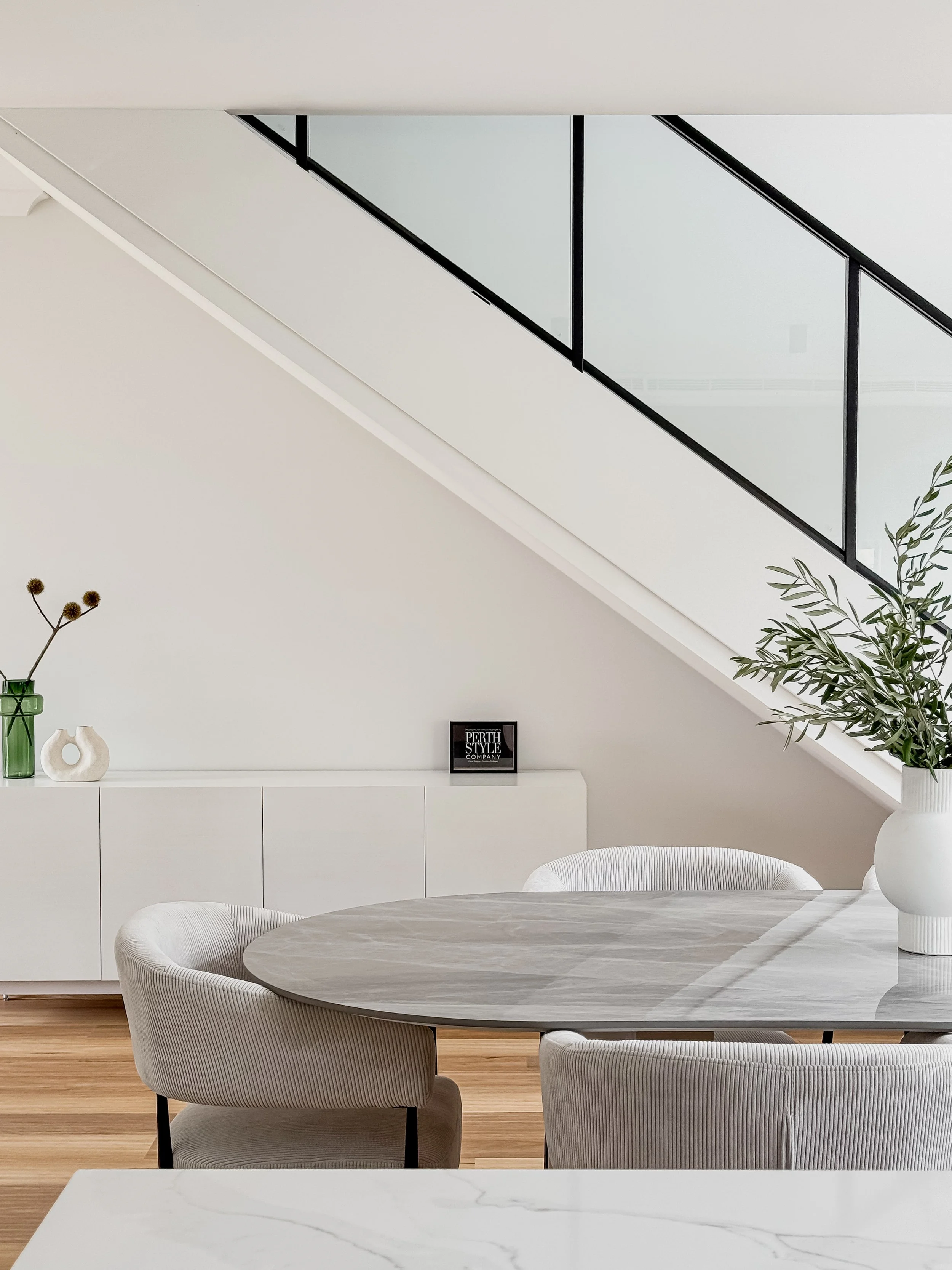 Modern dining area with a round marble table, white upholstered chairs, a white sideboard against the wall, decorative vases with plants, and a glass balcony railing above.