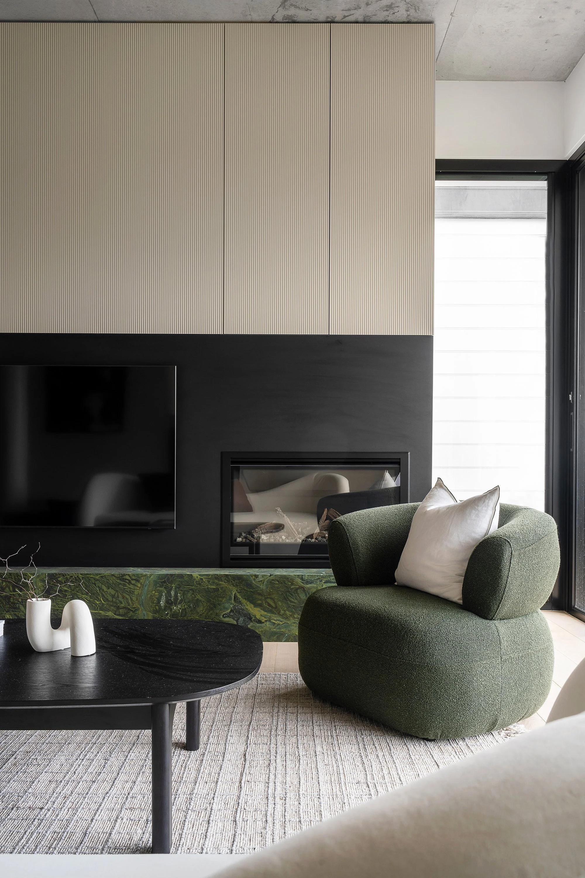 Modern living room with black and beige fireplace, green armchair with white pillows, black round coffee table with white vases, beige and gray patterned rug, large window with black frame, and beige cabinetry above the fireplace.