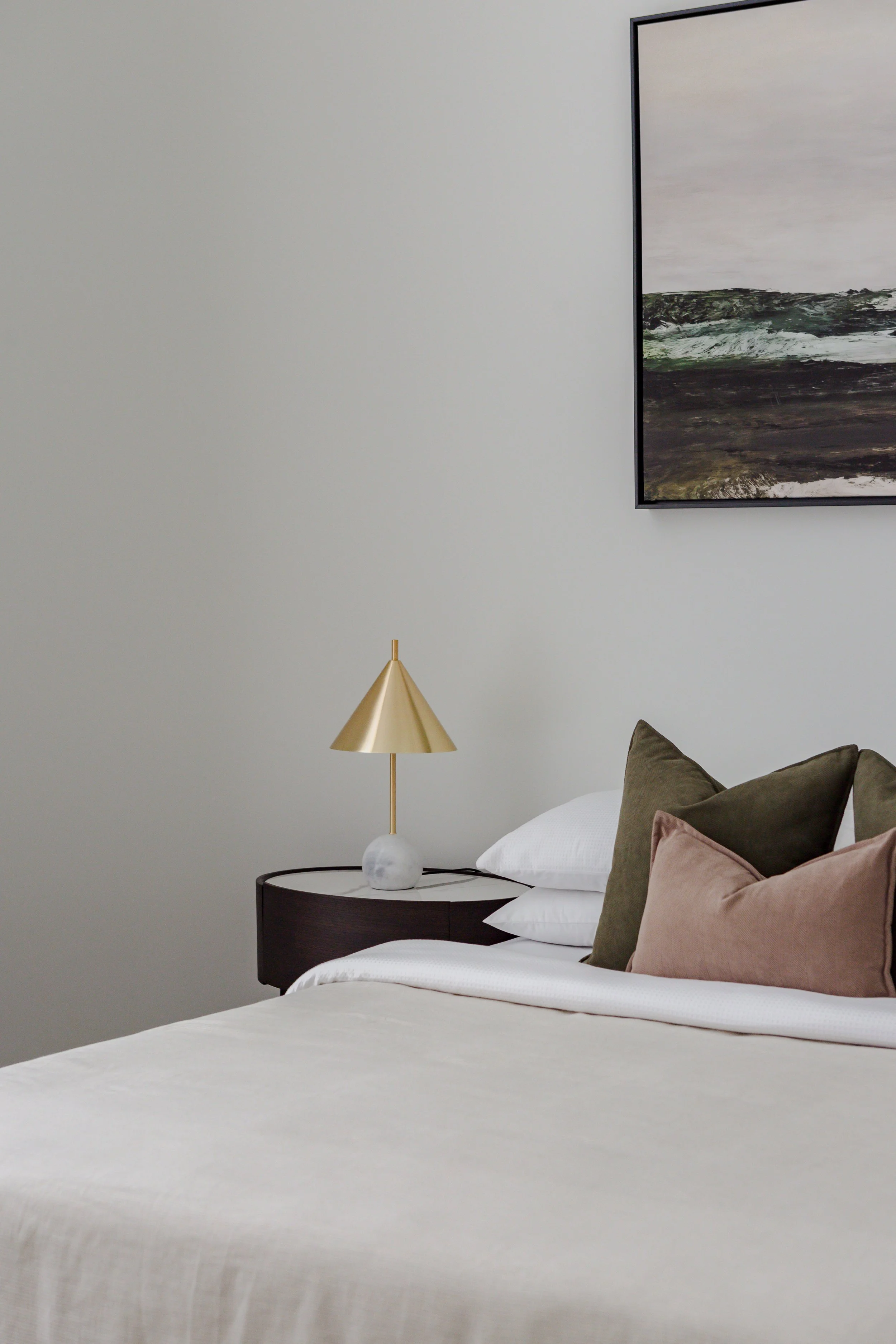 A neatly made bed with white sheets and multiple pillows, a black bedside table with a gold modern table lamp, and a framed abstract landscape painting on a plain light wall.