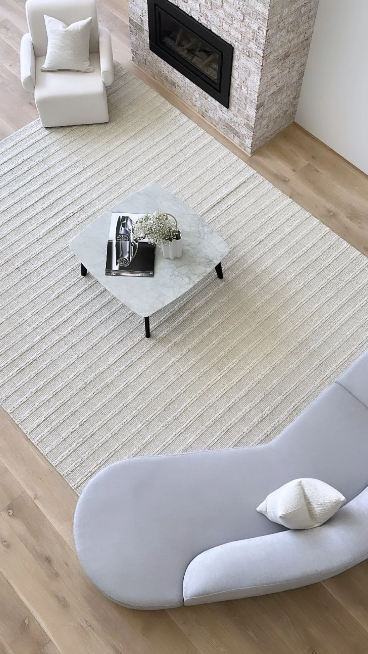 A modern living room with a beige striped rug, a marble-top coffee table, white armchairs, and a fireplace with a brick surround.